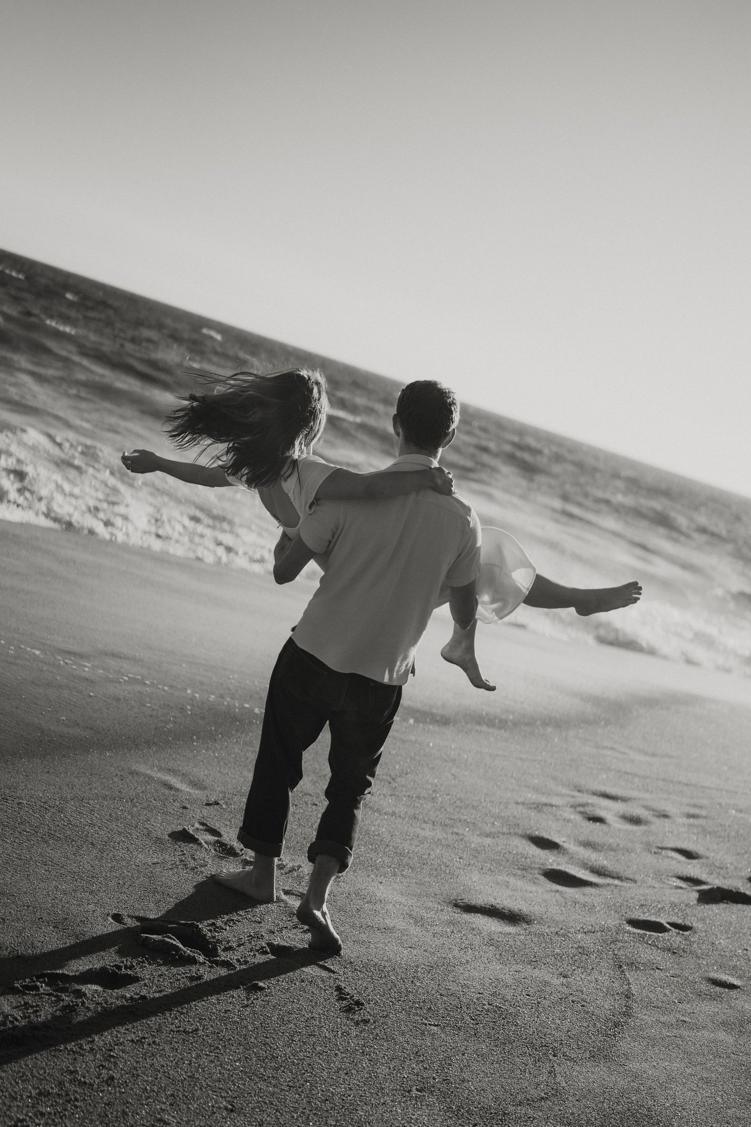 Black and white photo of a man carrying a woman on a sandy beach, footprints trailing behind them.