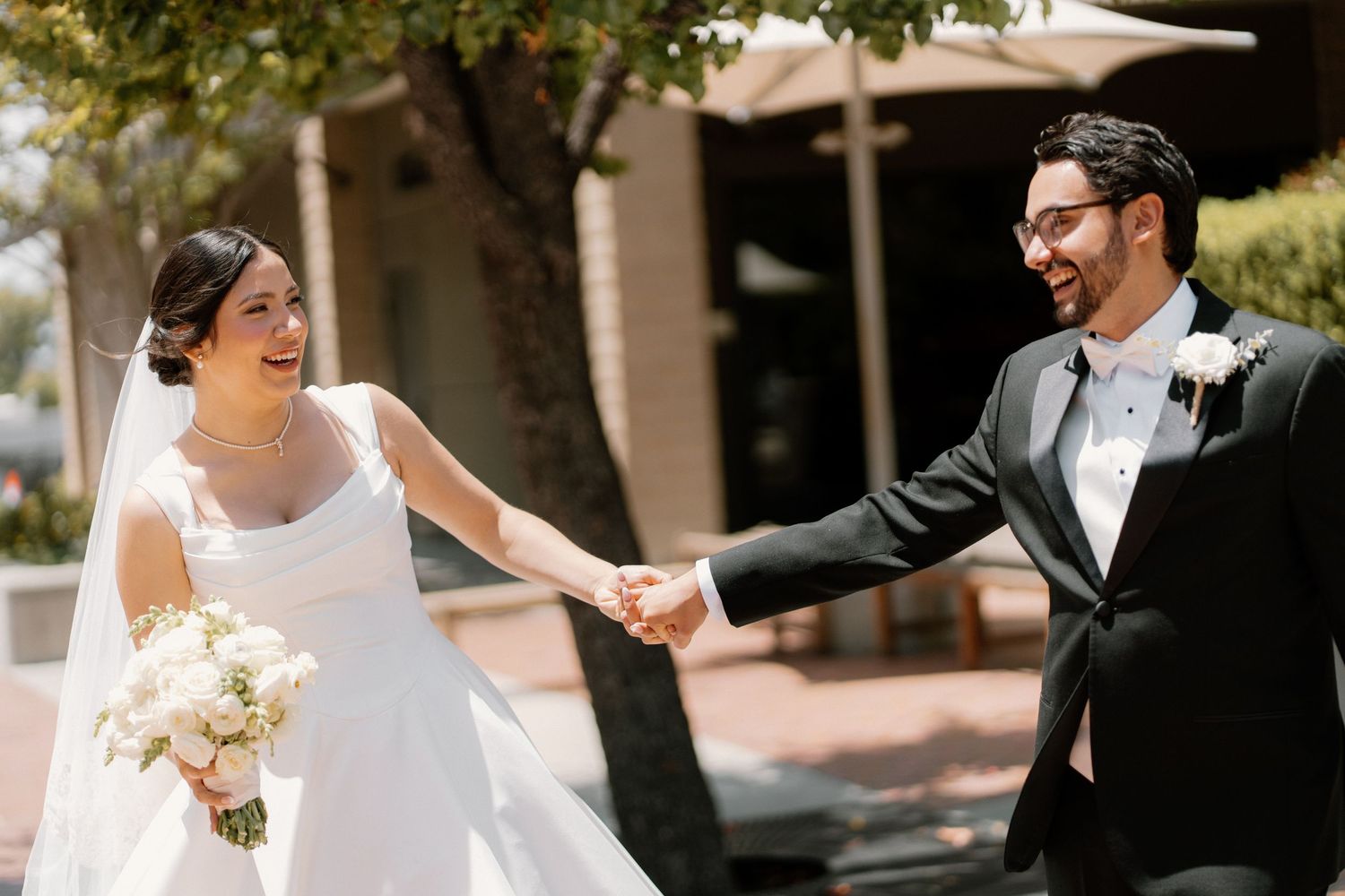 Smiling bride and groom hold hands outdoors on sunny wedding day.