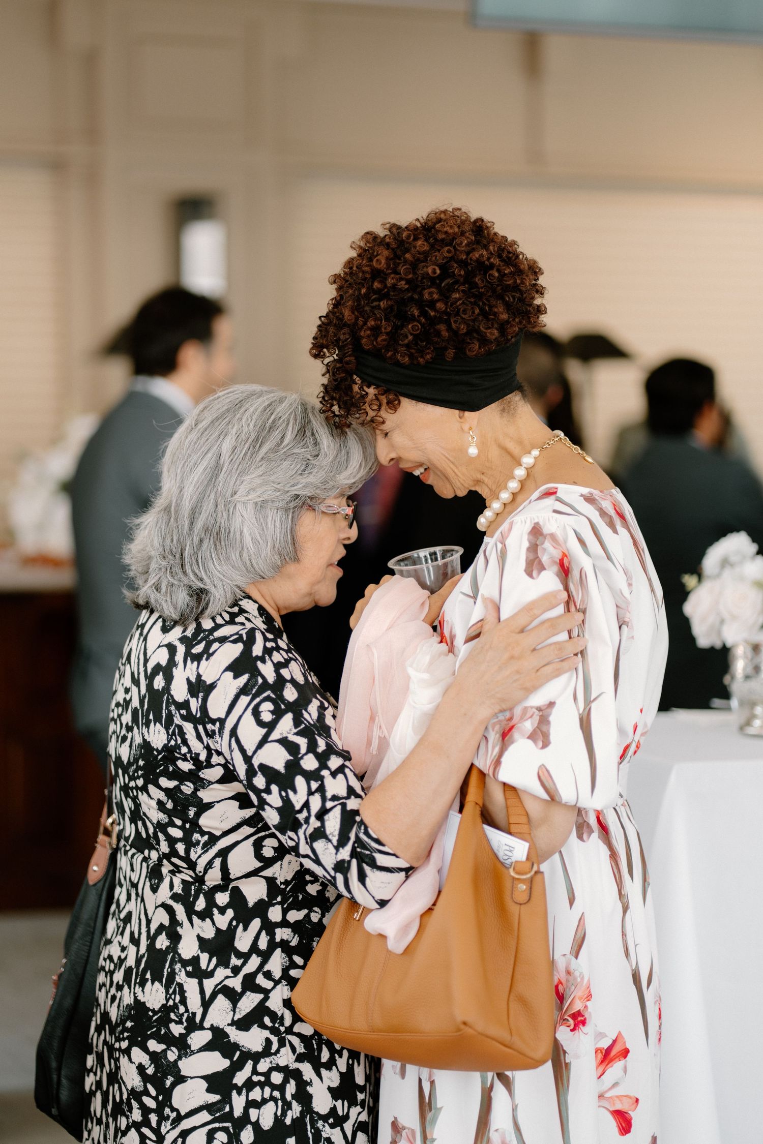 Two elegantly dressed women sharing a warm embrace and laugh at a wedding reception cocktail hour.