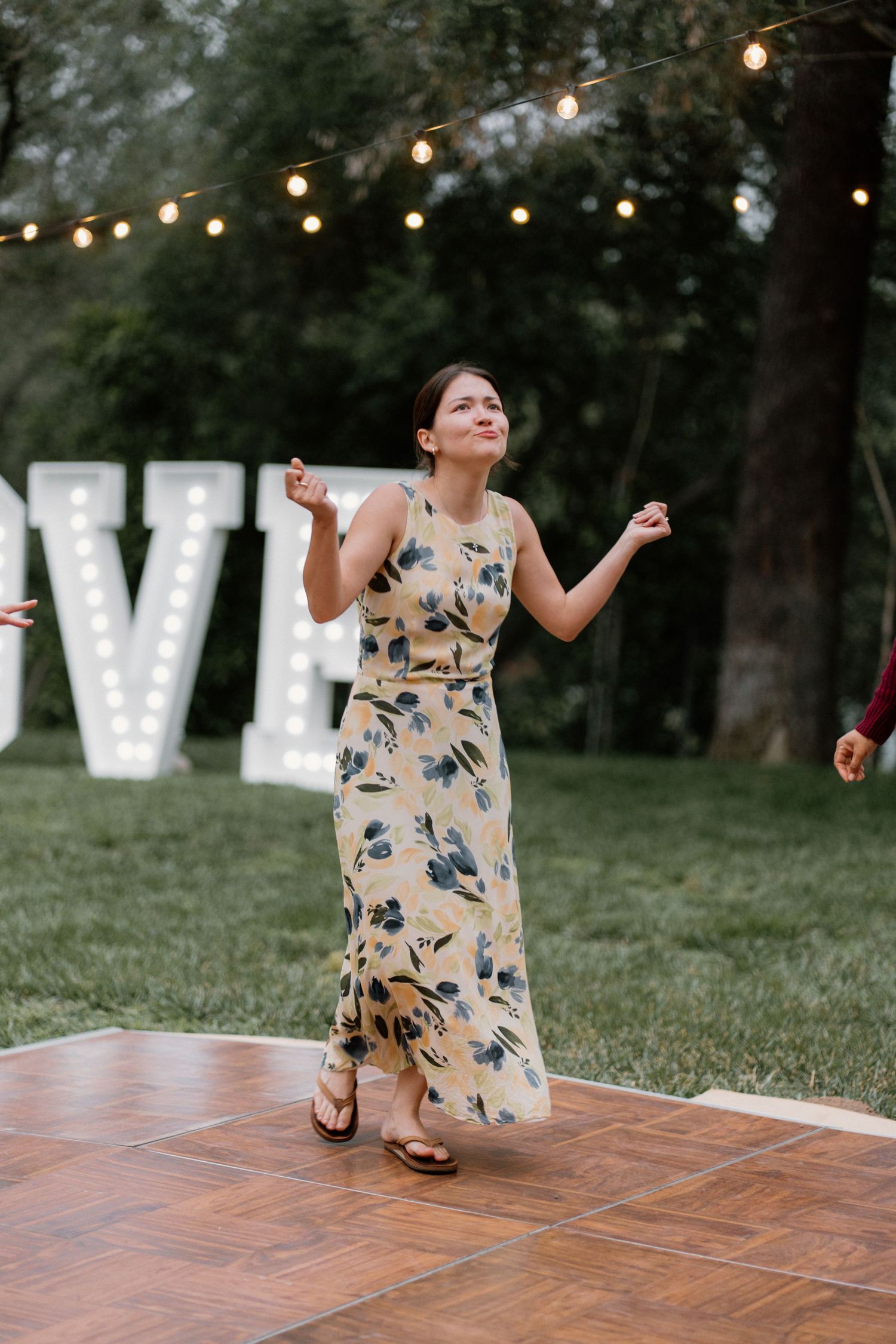 Woman in floral maxi dress dancing joyfully on wooden dance floor at outdoor wedding reception.