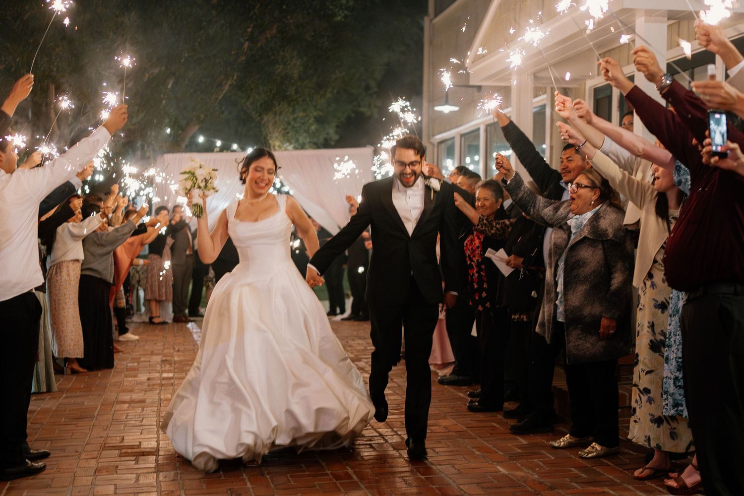 Newlyweds exit through sparkler tunnel surrounded by cheering guests at night wedding sendoff.