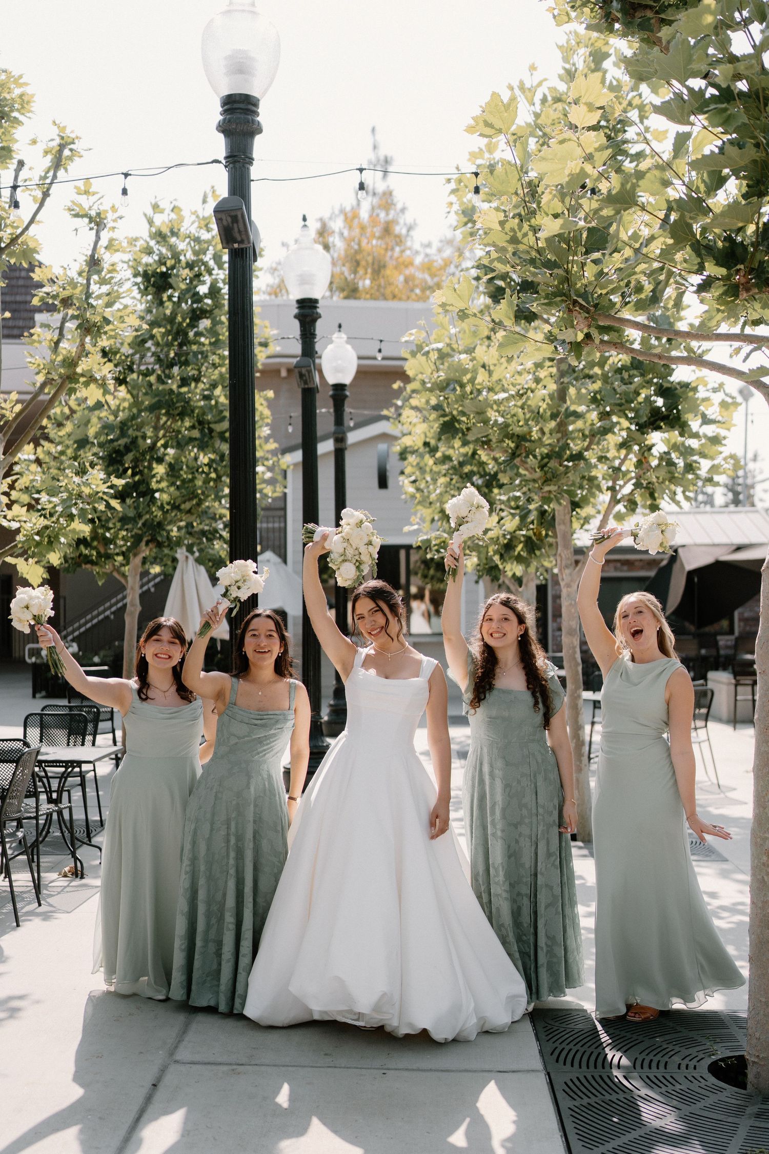 Bride in white gown celebrates with four bridesmaids in sage green dresses raising bouquets outdoors.