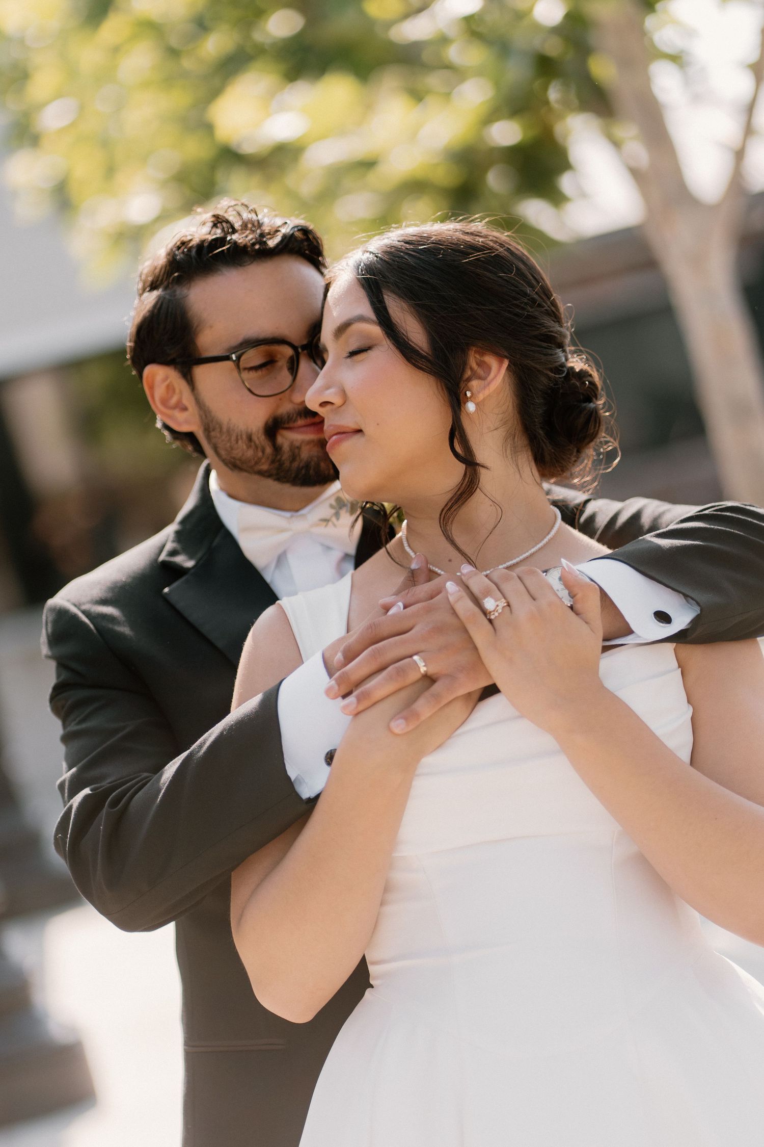 Groom in dark suit embraces bride in white gown from behind, both with eyes closed outdoors.