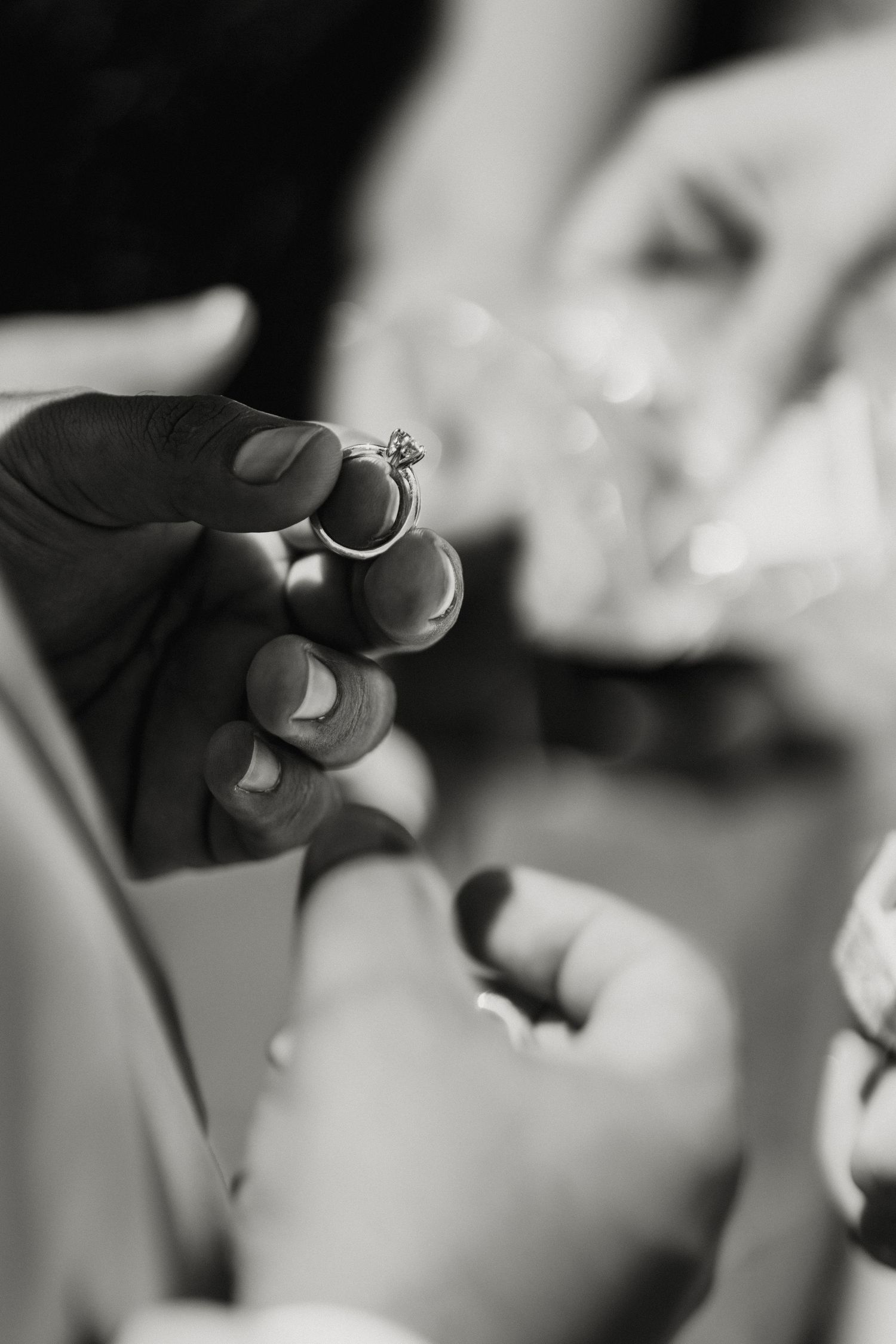 Black and white photo of hands holding a diamond engagement ring during a wedding proposal ceremony.