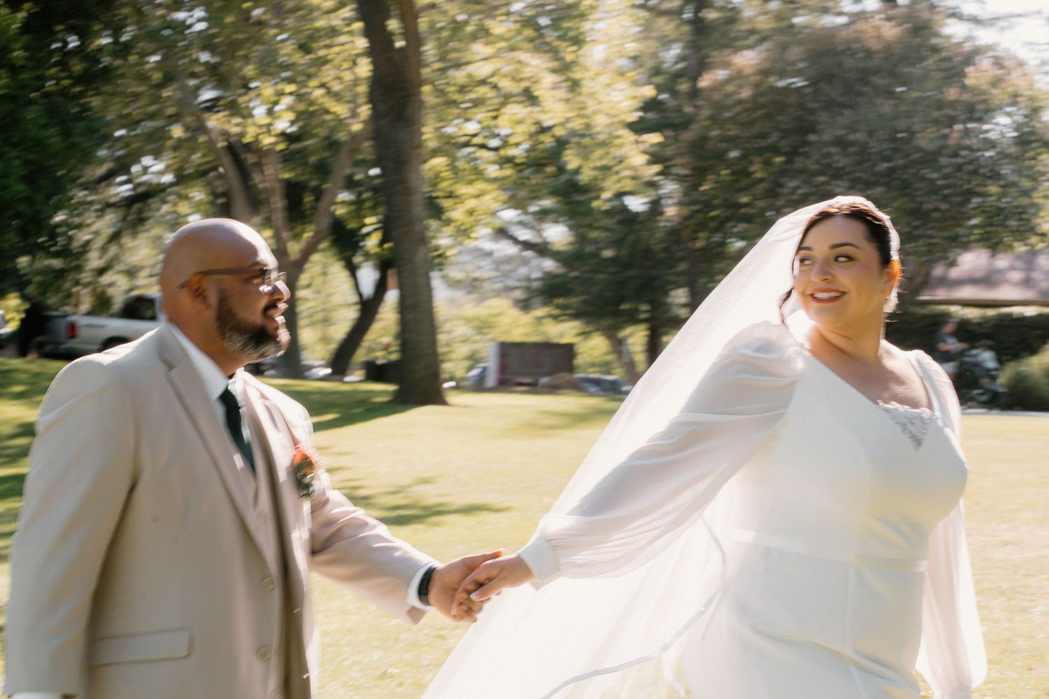Happy bride in white gown and veil holds hands with groom in tan suit walking in sunny park.