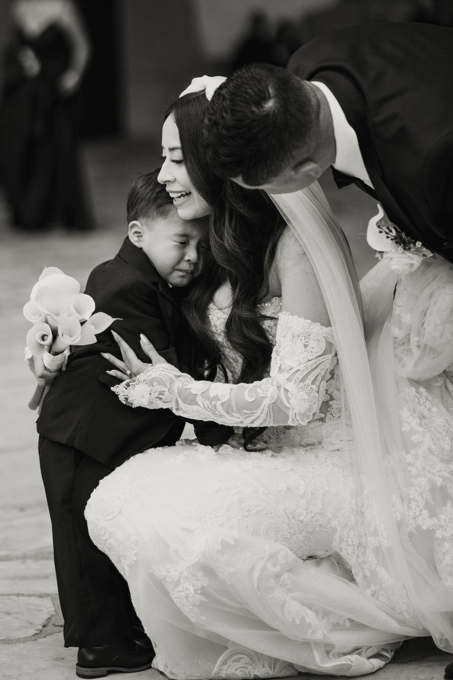 Black and white photo of a bride and groom sharing a tender moment hugging a young boy in a tuxedo.