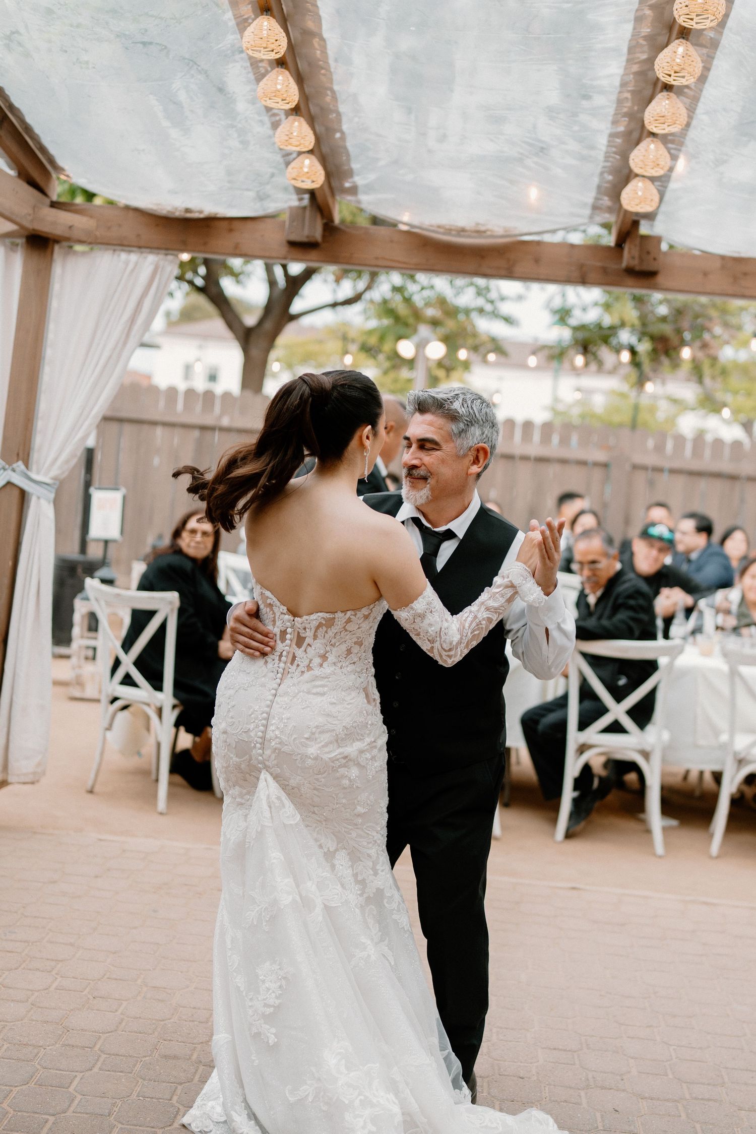 Bride in lace gown dancing with her father under a pergola at an outdoor wedding reception.