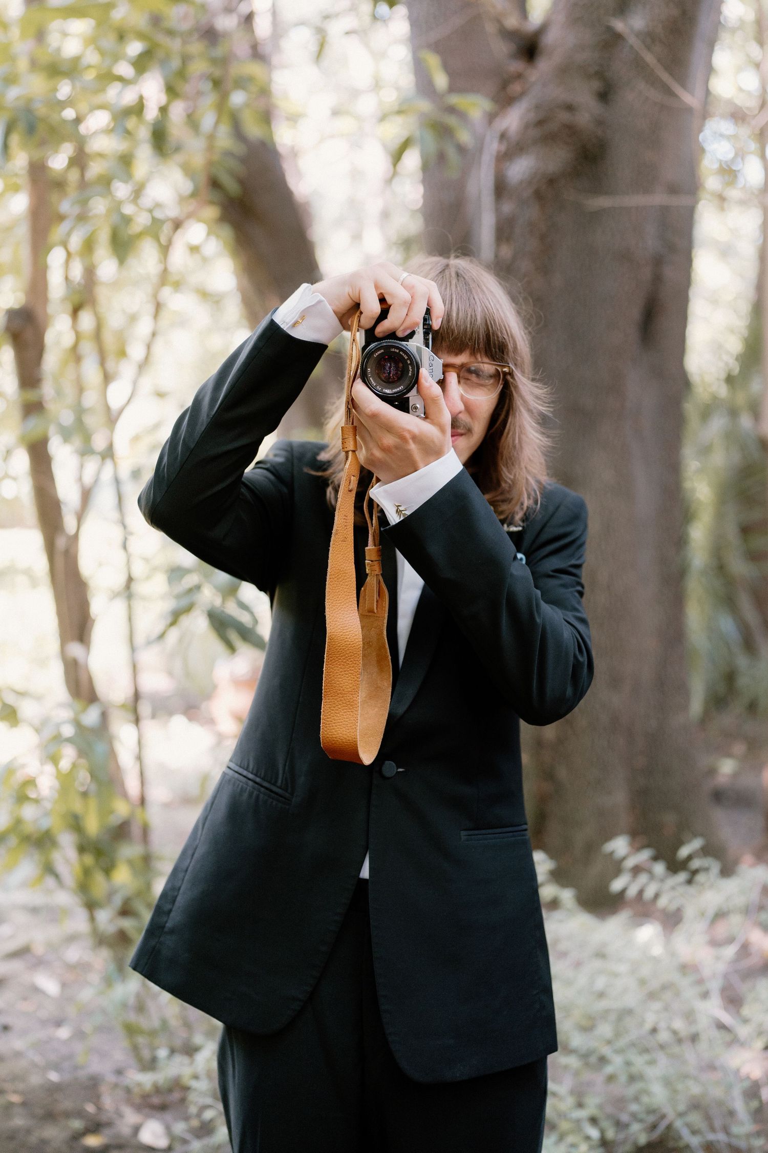 Photographer in black suit holding vintage camera up to take photo outdoors in wooded setting.