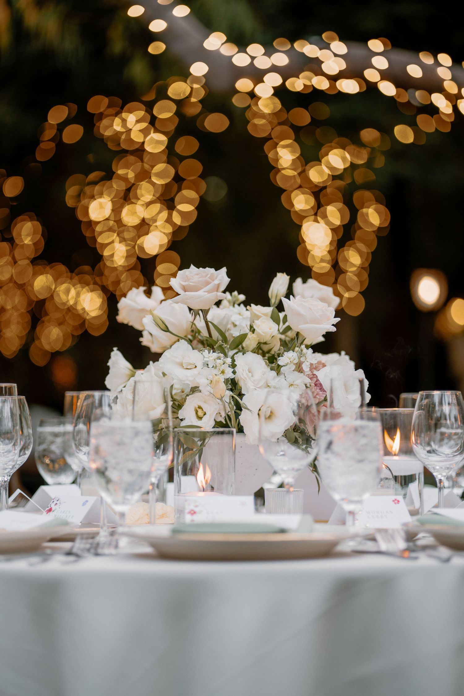 Elegant wedding table setting with white roses, candles, crystal glassware, and glowing bokeh string lights.