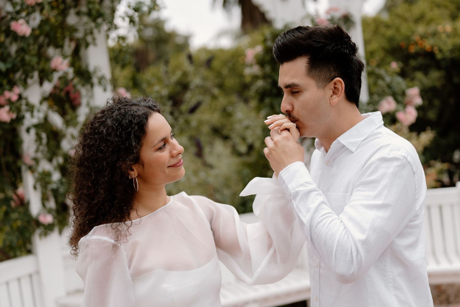 A man in a white shirt gently kisses a woman's hand as she smiles at him outdoors.