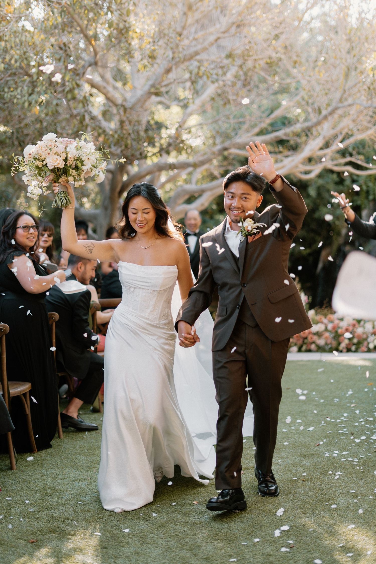 Joyful newlywed couple walks down outdoor aisle as guests toss white flower petals at their wedding ceremony.