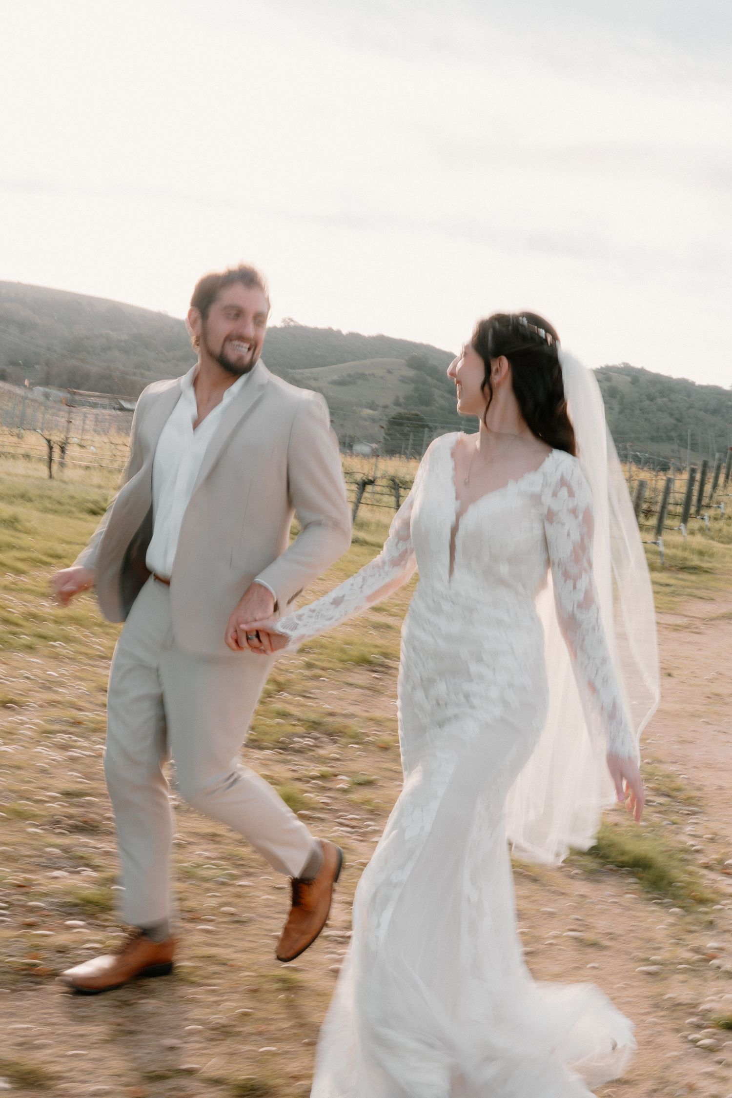 Laughing bride and groom running hand-in-hand through vineyard field at golden hour.