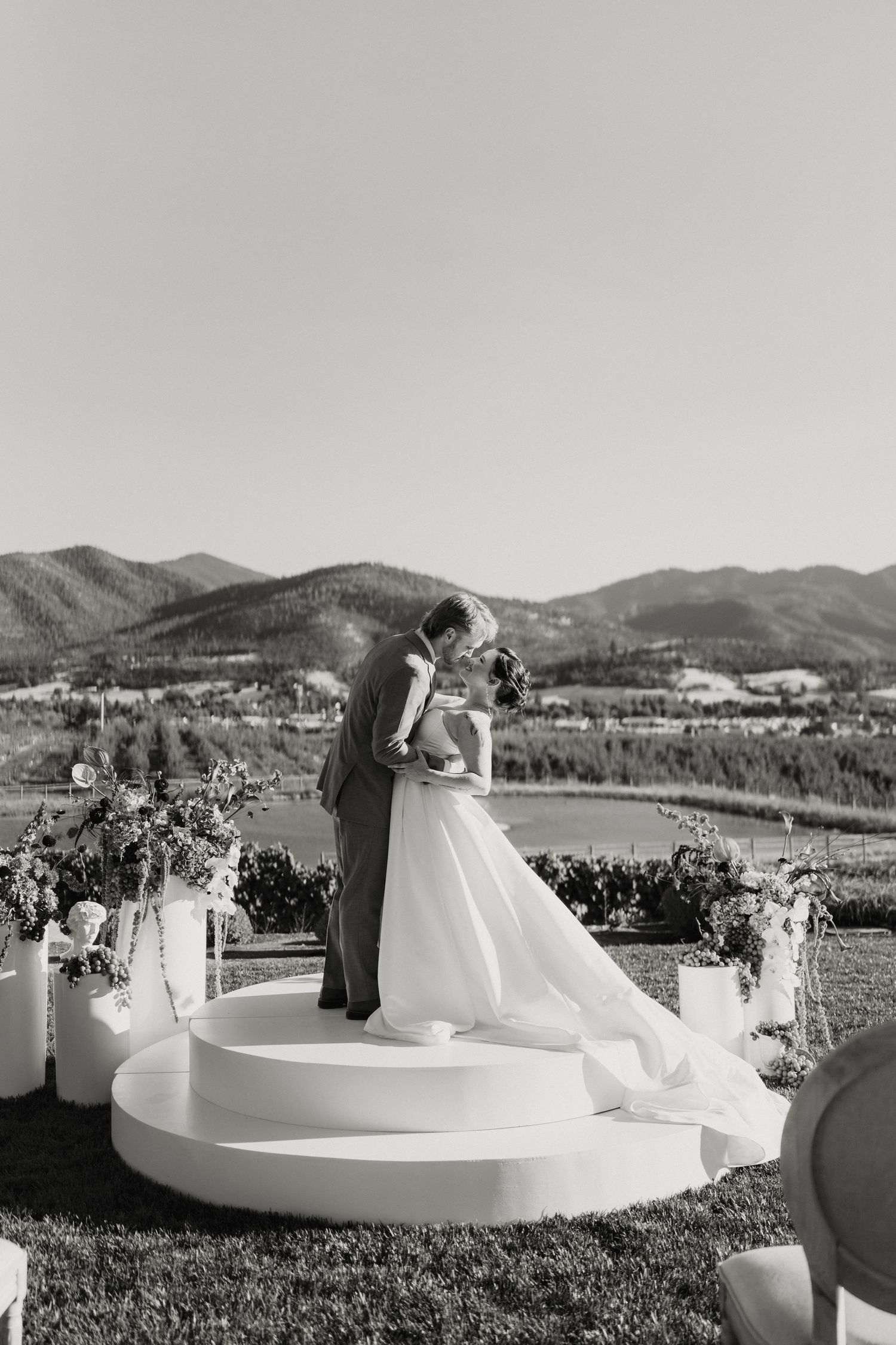 Black and white wedding photo of bride and groom kissing on circular altar with mountain lake backdrop.