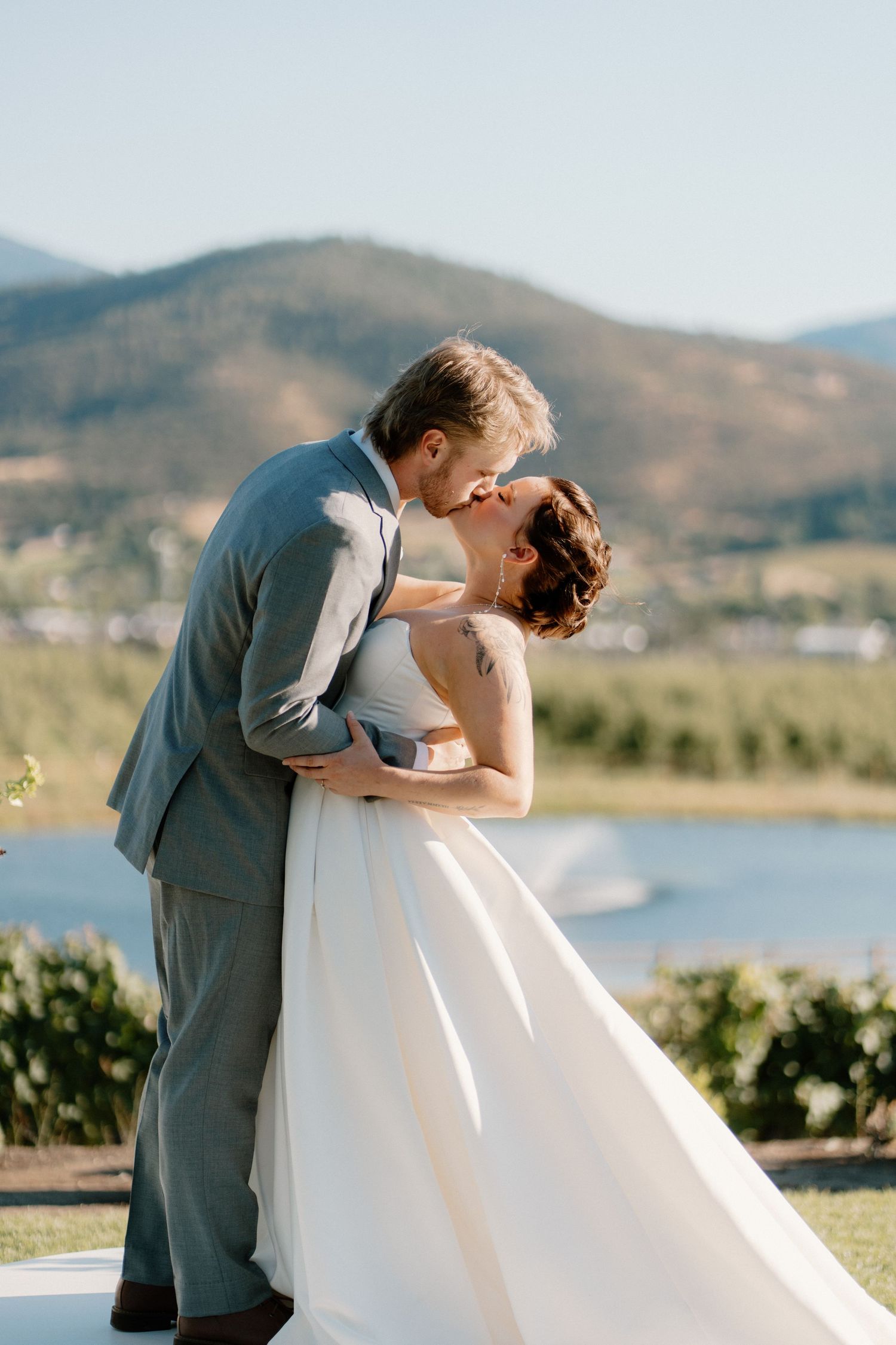 Bride and groom share first kiss at outdoor wedding ceremony with scenic mountain lake backdrop on sunny day.