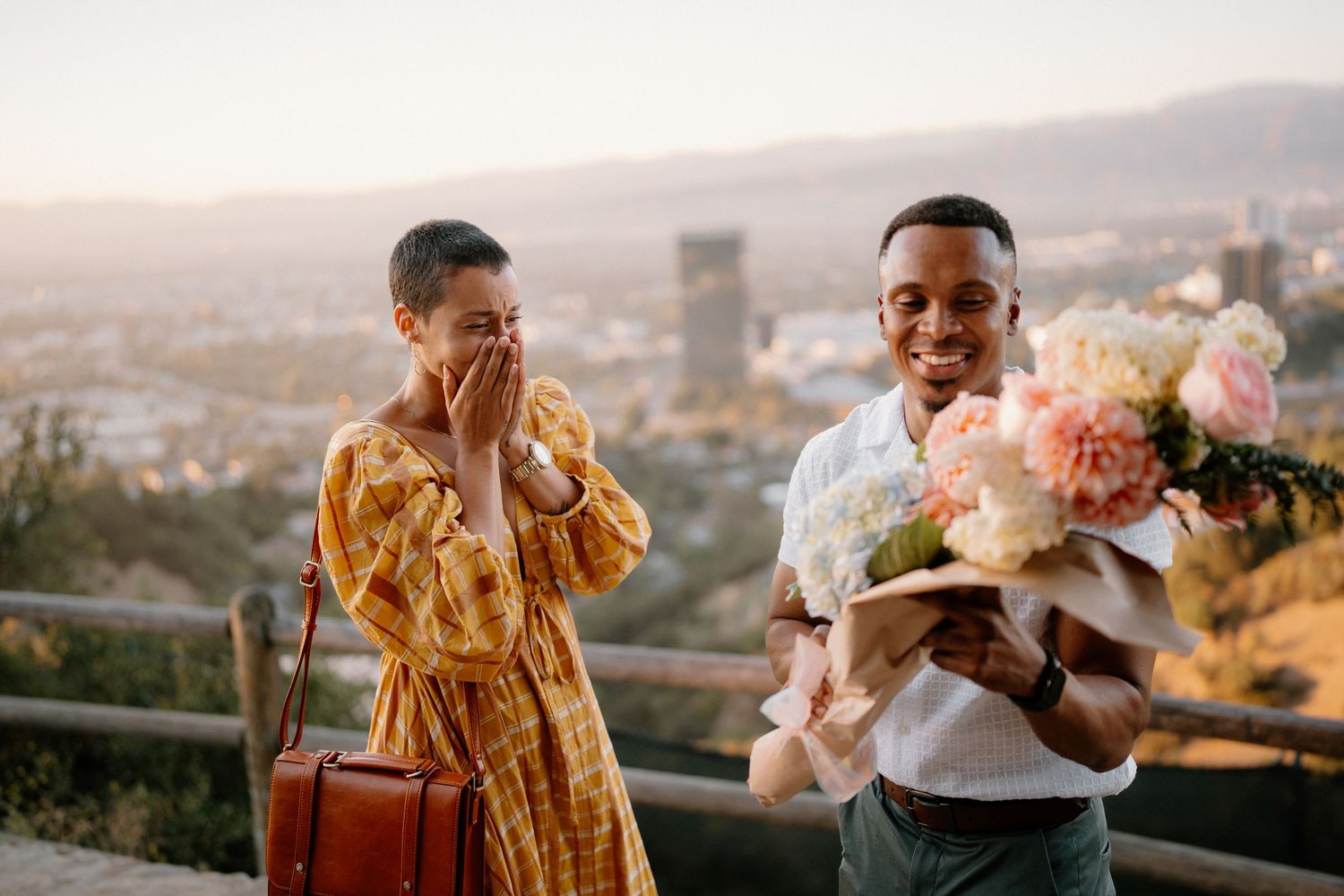 A man surprises a woman with a bouquet of flowers at a scenic overlook during golden hour.