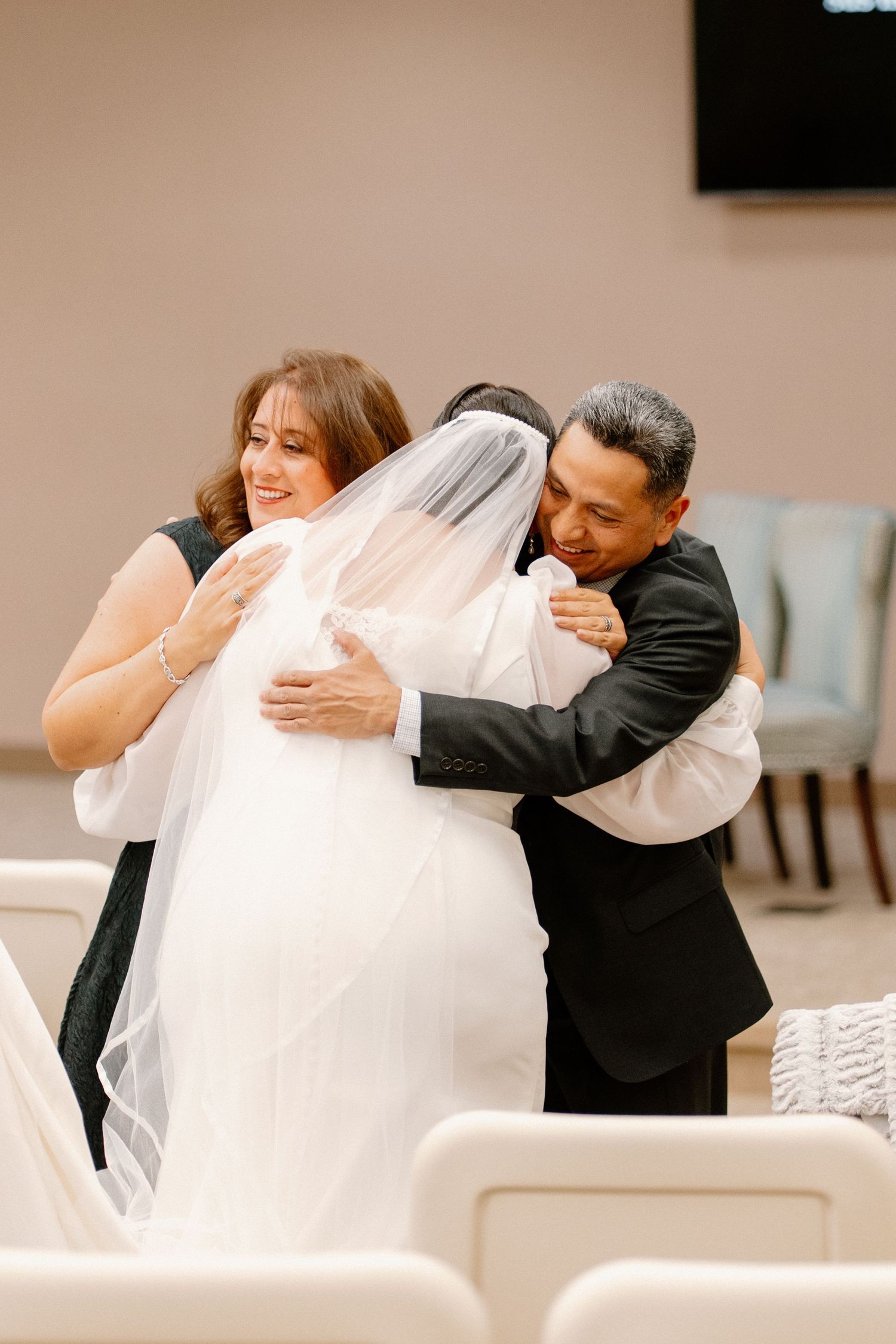 Bride in white veil shares emotional group hug with parents in black attire during wedding ceremony.