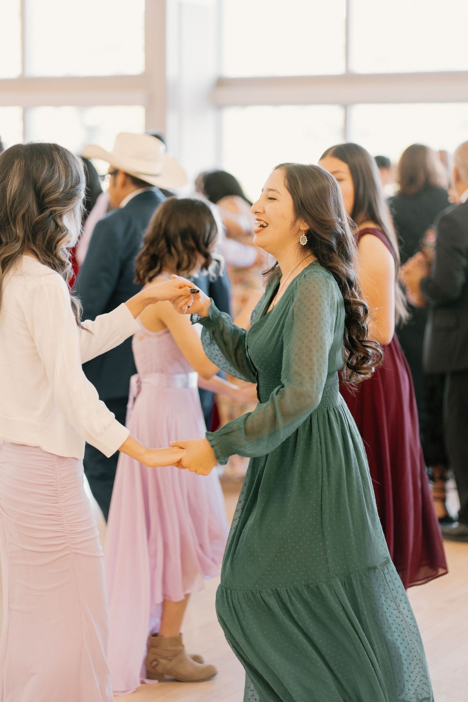Women in green and pink dresses laughing and dancing together at a lively wedding reception.