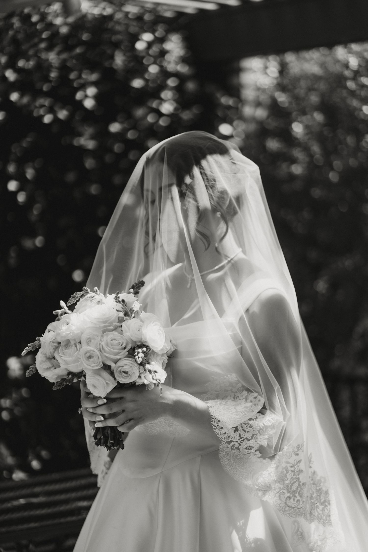 Black and white portrait of bride in lace gown holding white rose bouquet with veil draped over face.