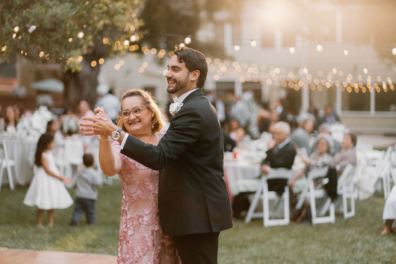 Groom dances with his mother at an outdoor wedding reception under warm string lights at dusk.