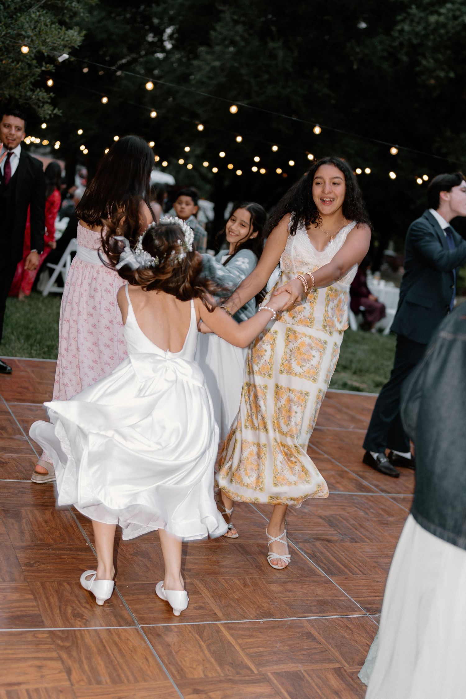 Bride and guests dancing joyfully on outdoor dance floor with string lights at evening wedding reception.