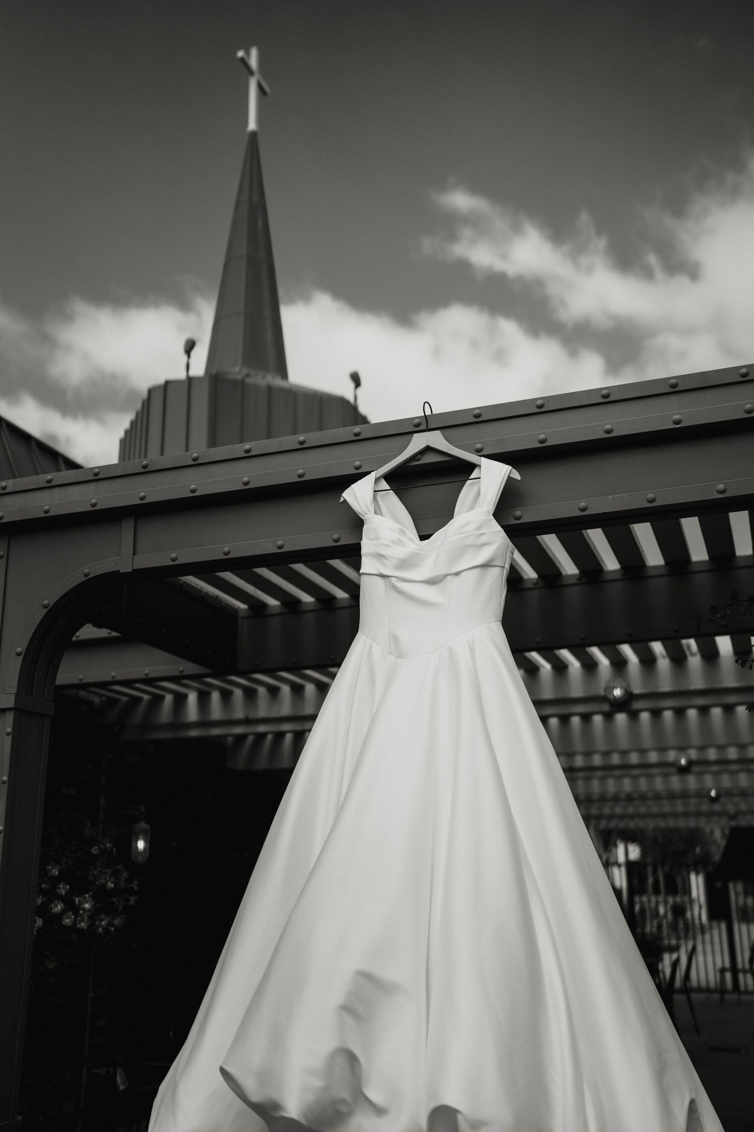 Black and white photo of an elegant wedding gown hanging on a hanger outside a church with a steeple.