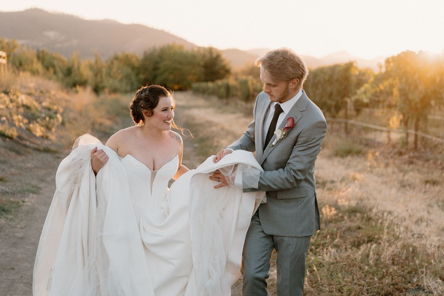 A groom in a gray suit helps his bride with her white wedding gown during a golden hour outdoor photo session.