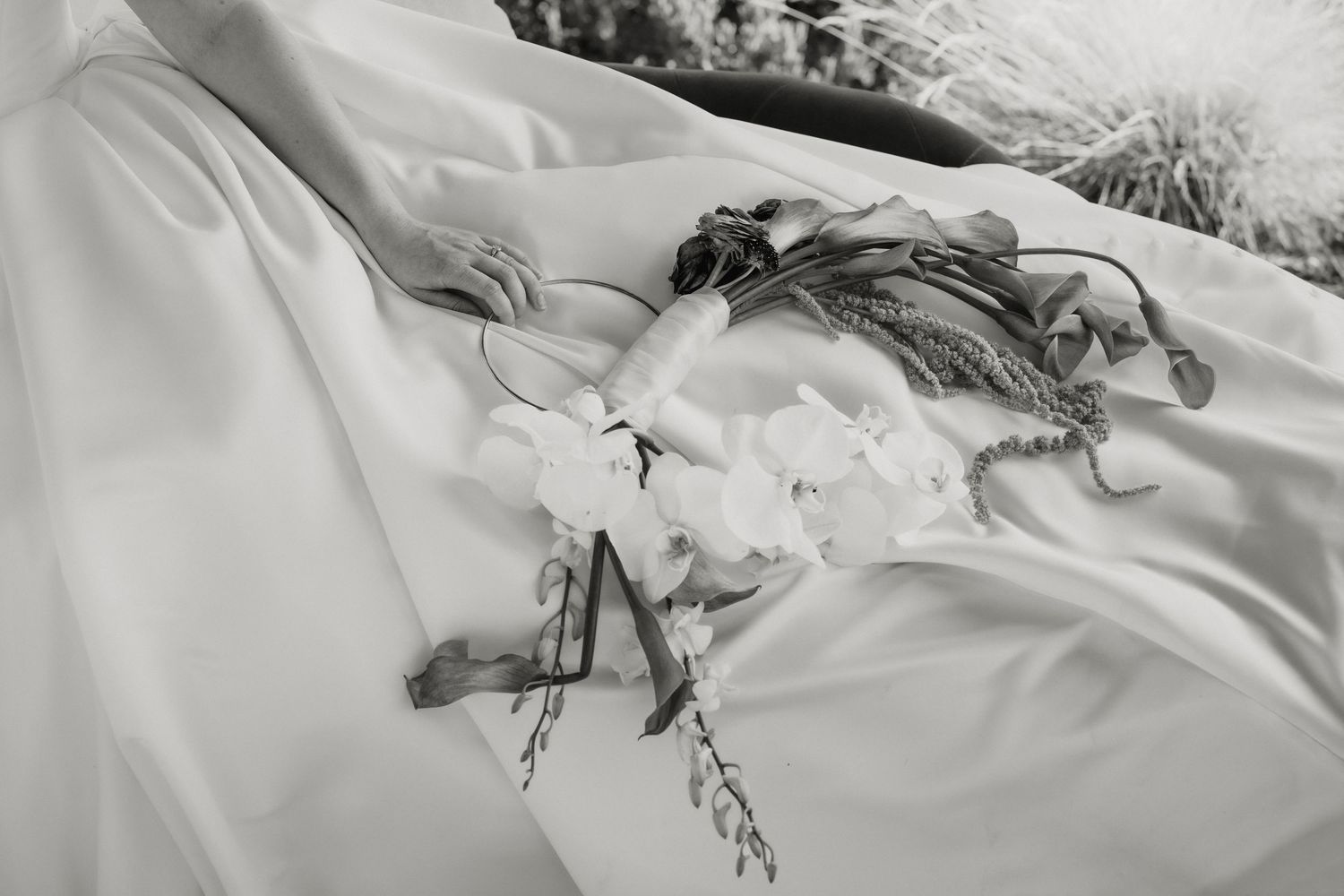 Black and white photo of a bridal bouquet with white orchids and greenery resting on a flowing white wedding dress.