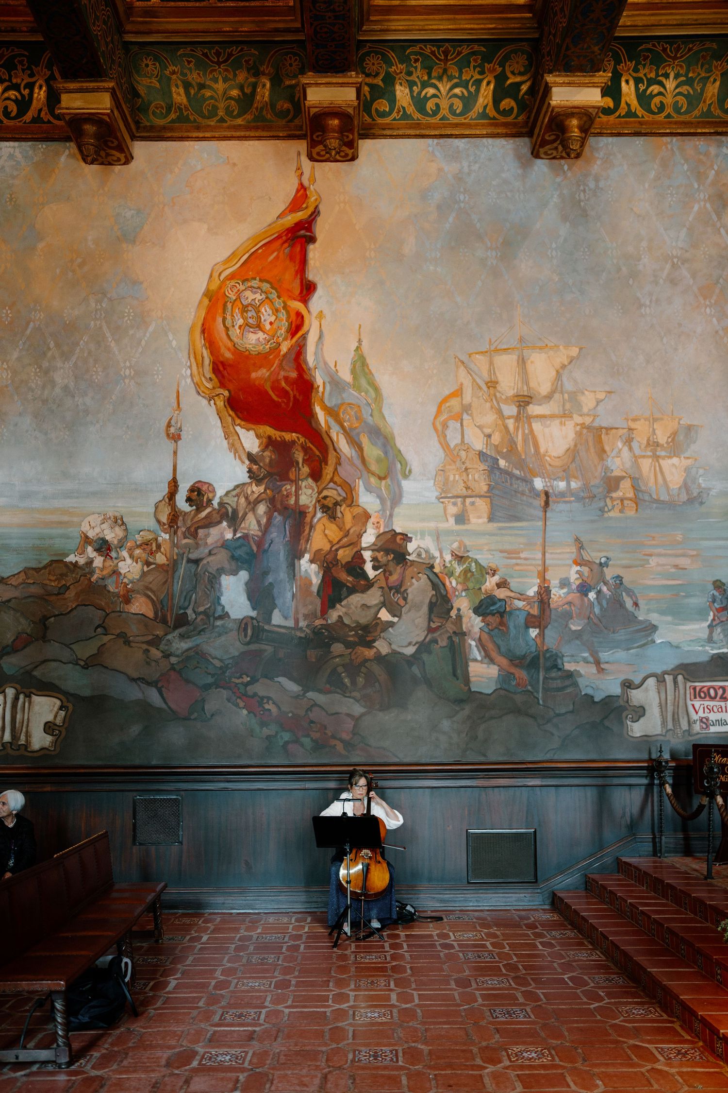 Cellist performing beneath large historic mural painting in elegant wedding ceremony venue.
