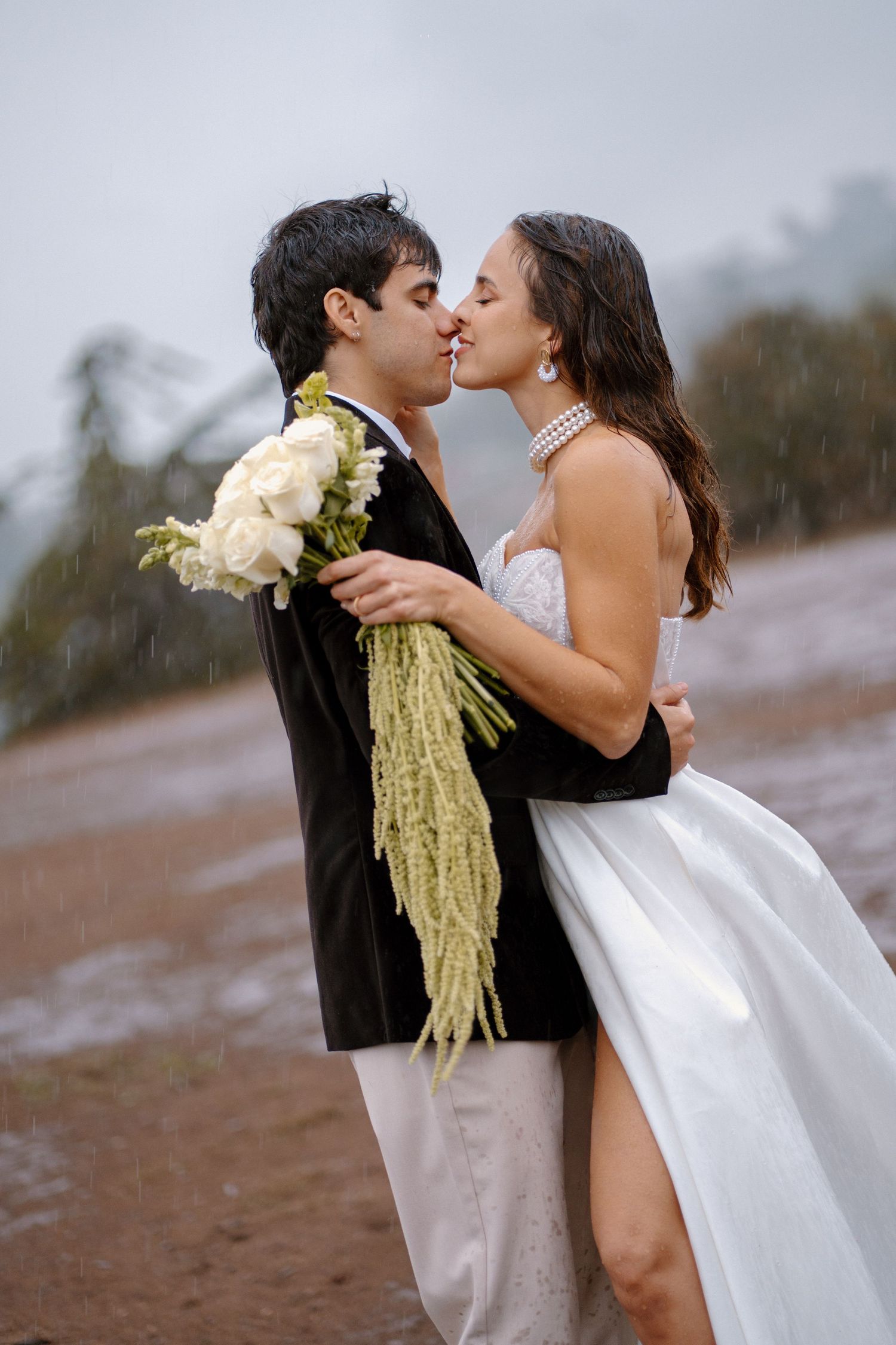 Newlyweds share a romantic kiss in the rain, bride in strapless white gown holding a white and green bouquet.