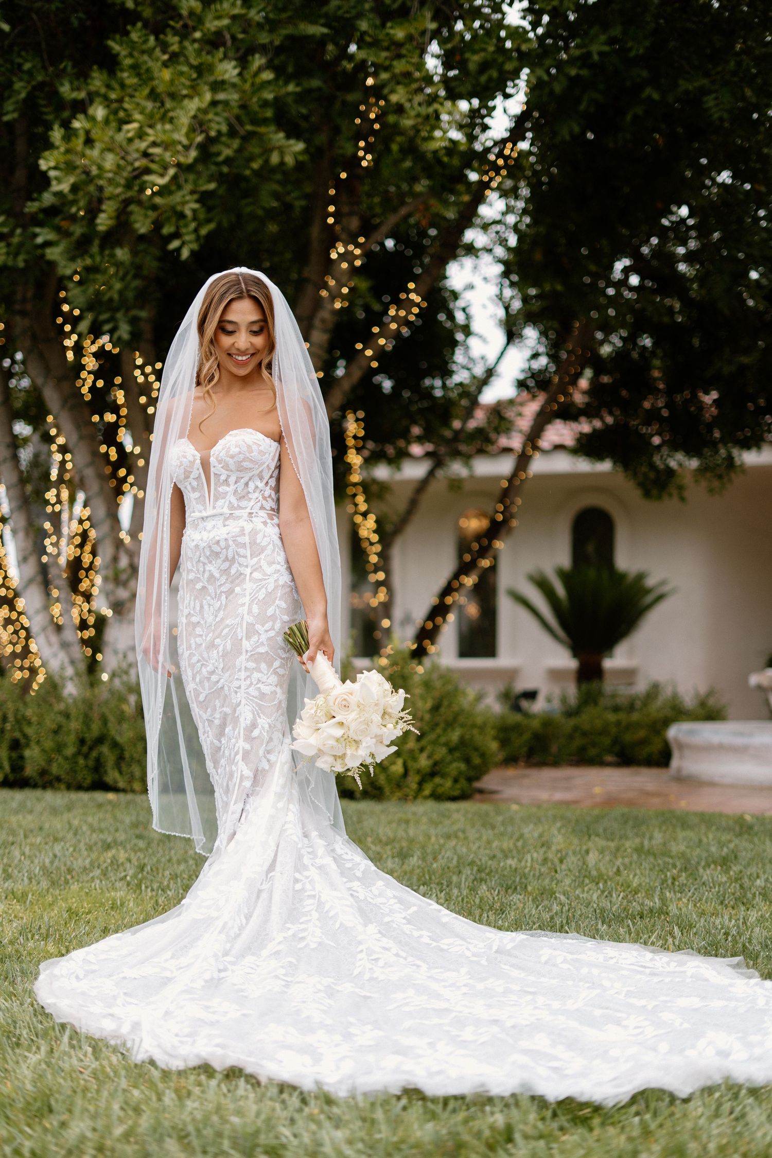 Beautiful bride in a fitted lace wedding gown with long veil holding white bouquet in a garden with fairy lights.