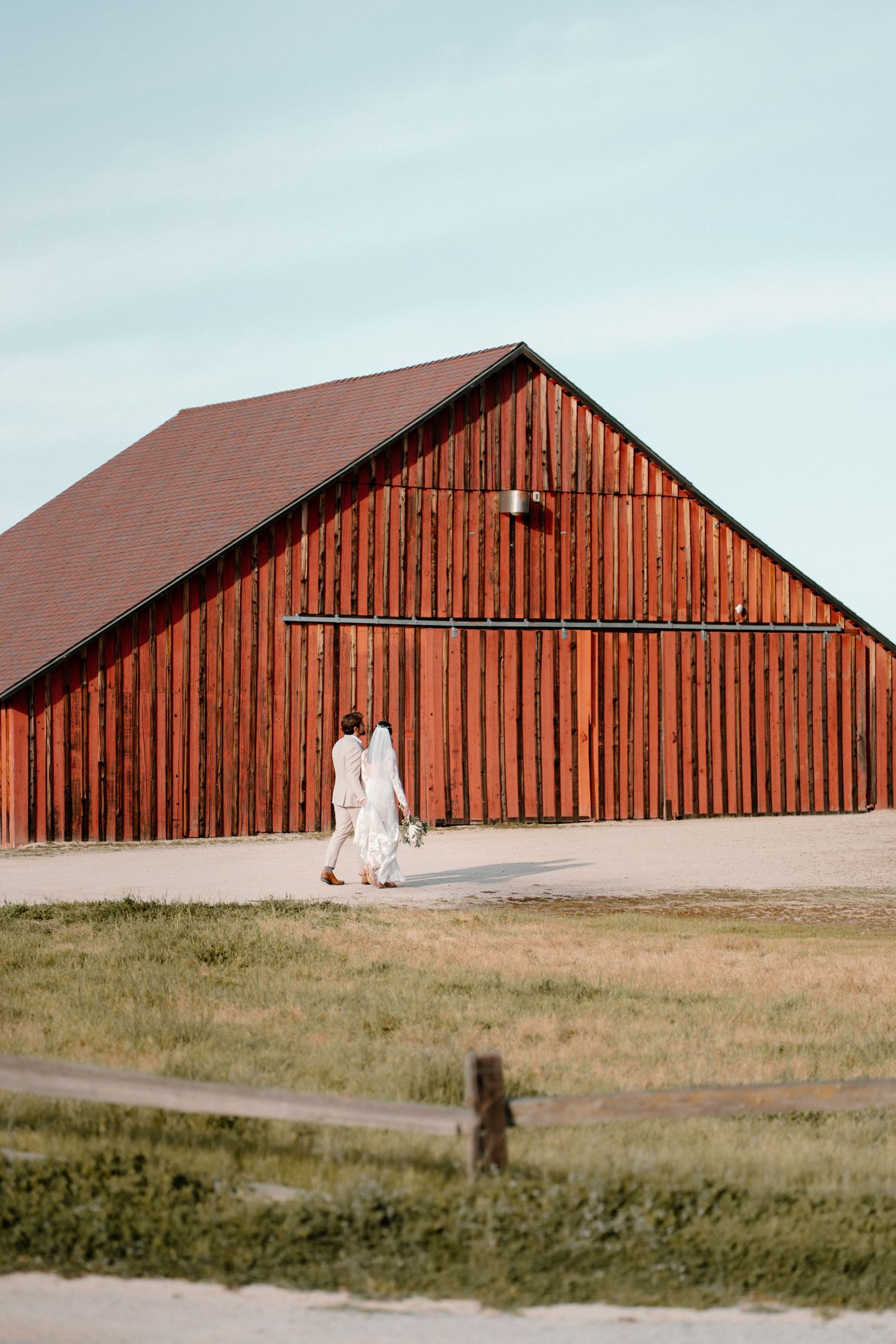 Bride and groom walking hand in hand past a large rustic red barn on their wedding day.