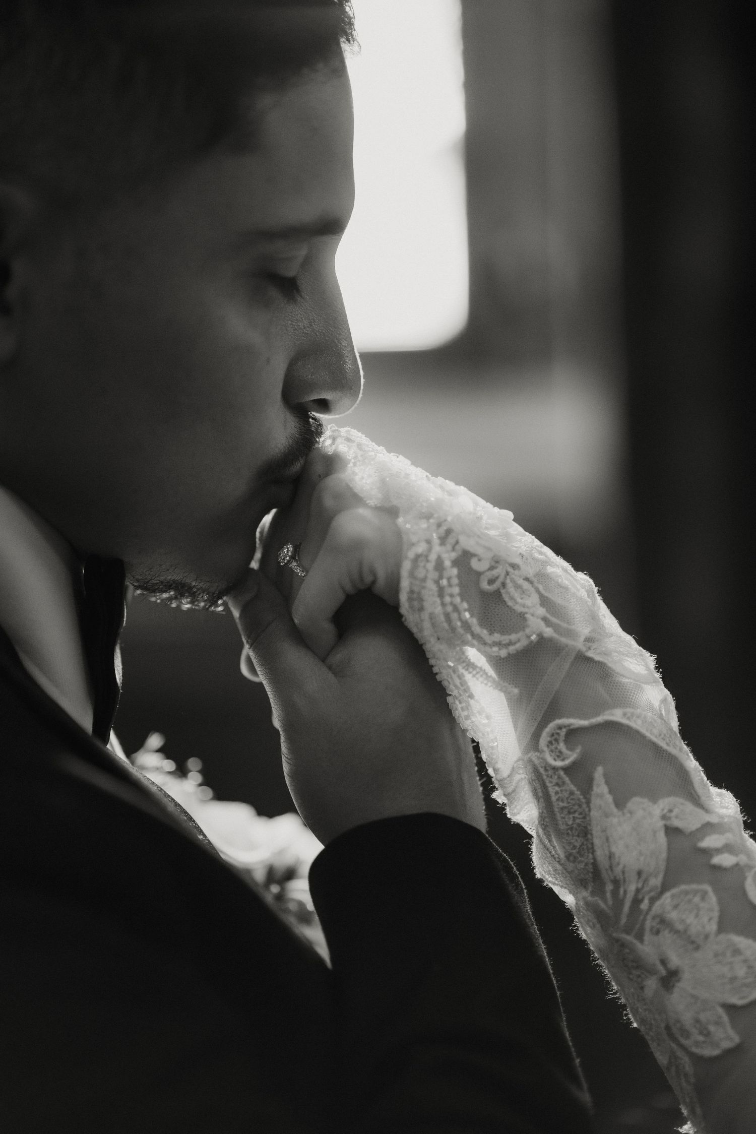 Groom kissing bride's lace-gloved hand in romantic black and white wedding portrait.
