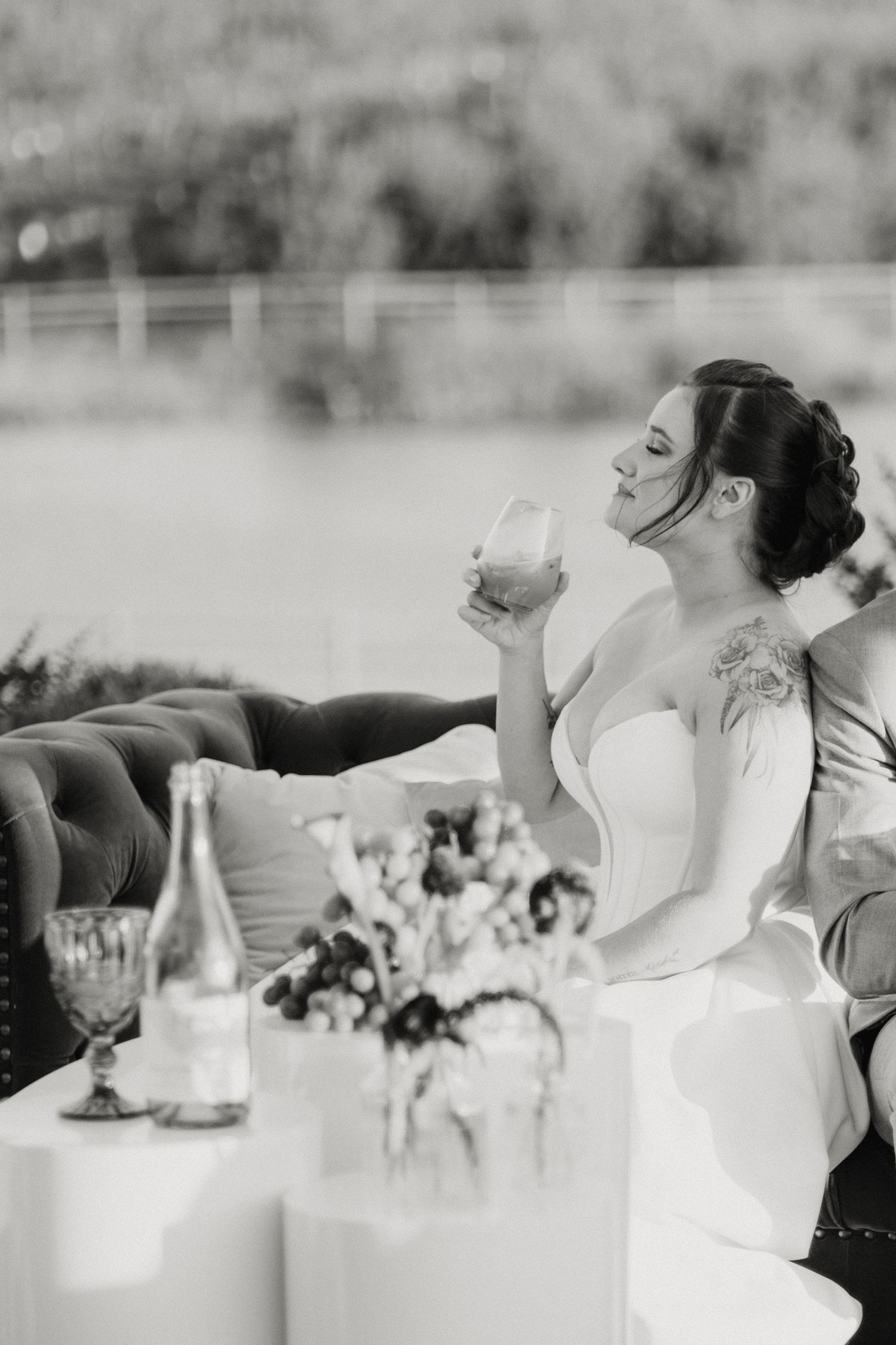 Black and white photo of a bride in a strapless gown relaxing on a sofa, sipping from a glass at her wedding reception.