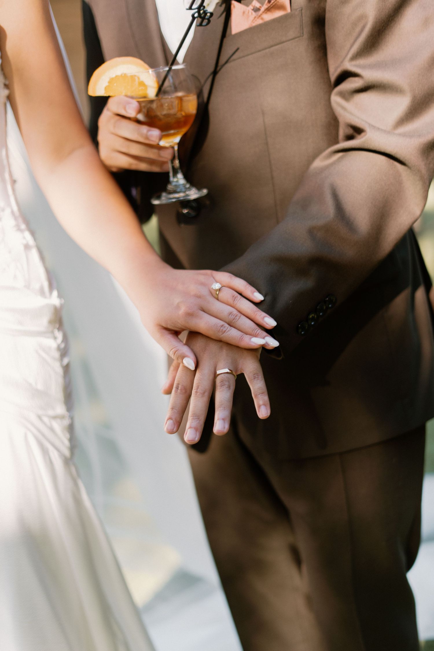 Close-up of bride and groom showing off gold wedding rings while holding a cocktail at their reception.