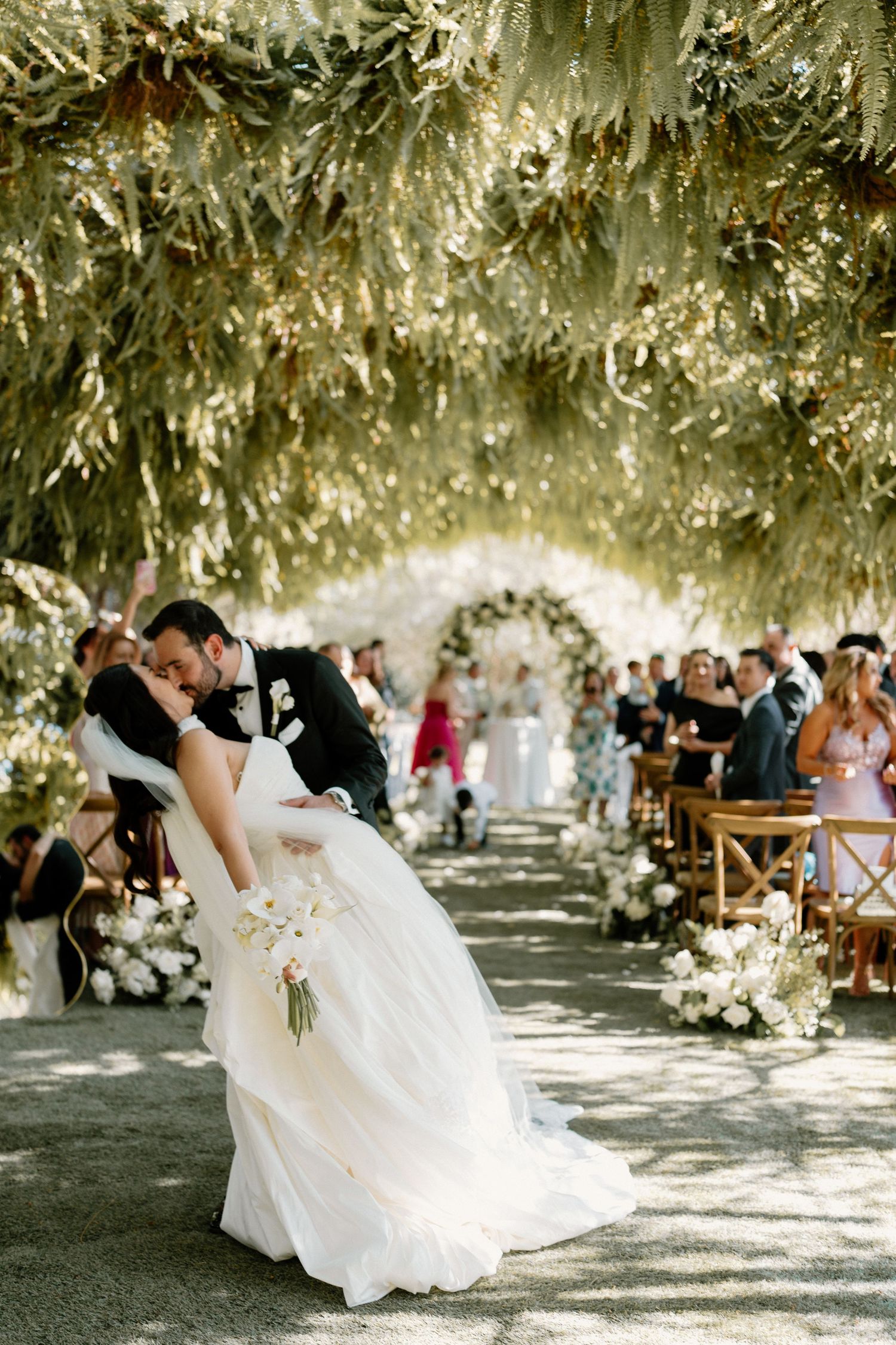 A groom dips his bride for a kiss along a floral-lined aisle under a lush green canopy.