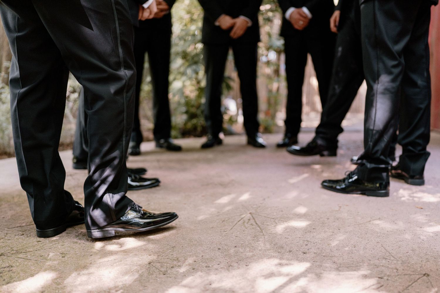 Close-up of groomsmen in black suits and polished dress shoes standing in a circle outdoors.