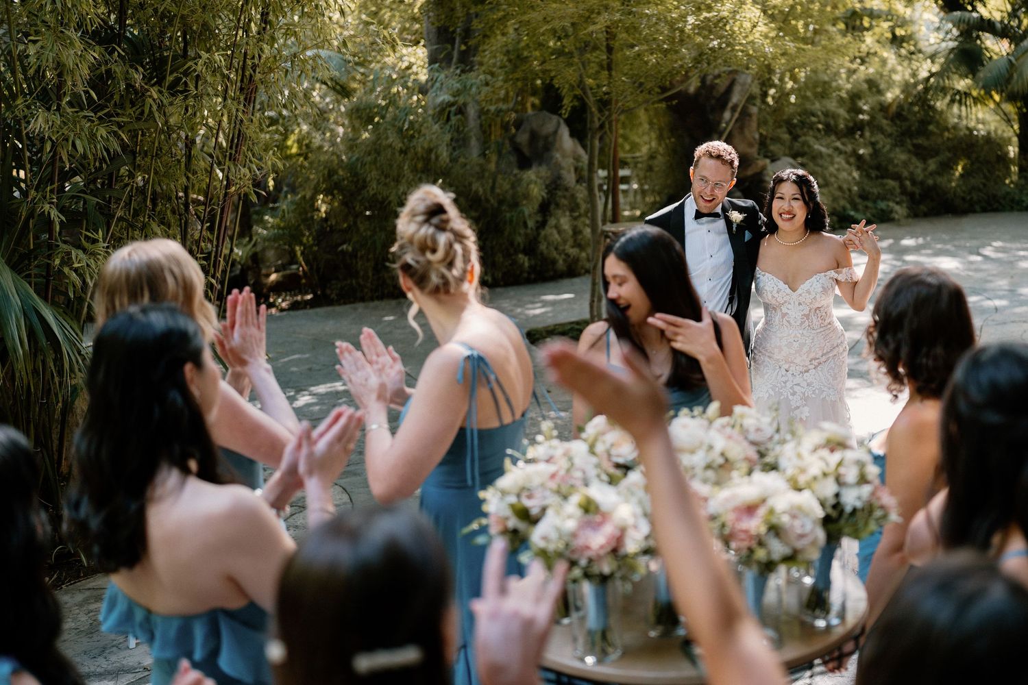 Bride and groom arrive to cheering bridesmaids in blue dresses at an outdoor garden wedding ceremony.