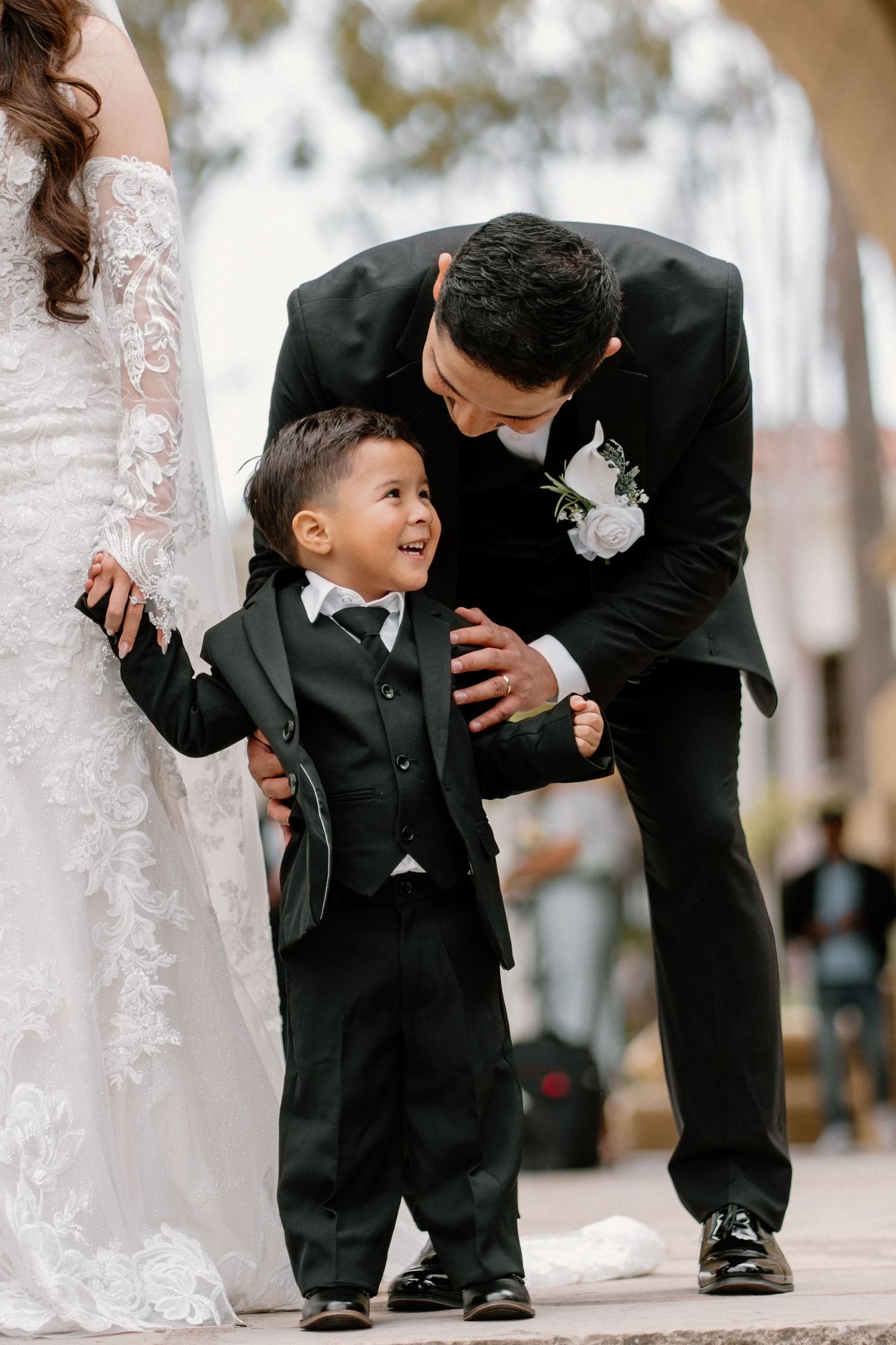 A groom in a black suit lovingly leans down to interact with a smiling young boy in a matching tuxedo.