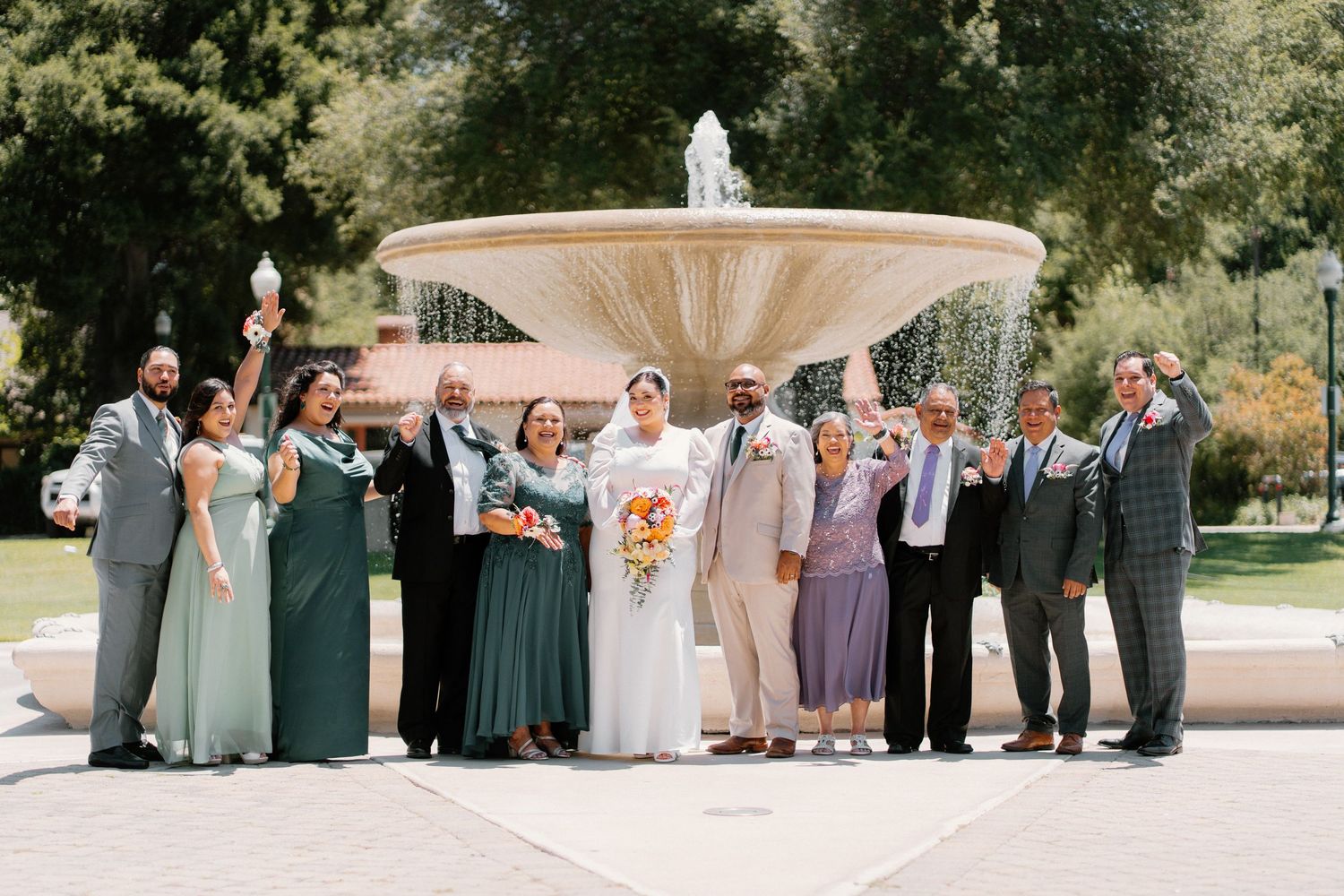 Wedding party poses by ornate fountain outdoors, bride in white gown holding bouquet, bridesmaids in green dresses.