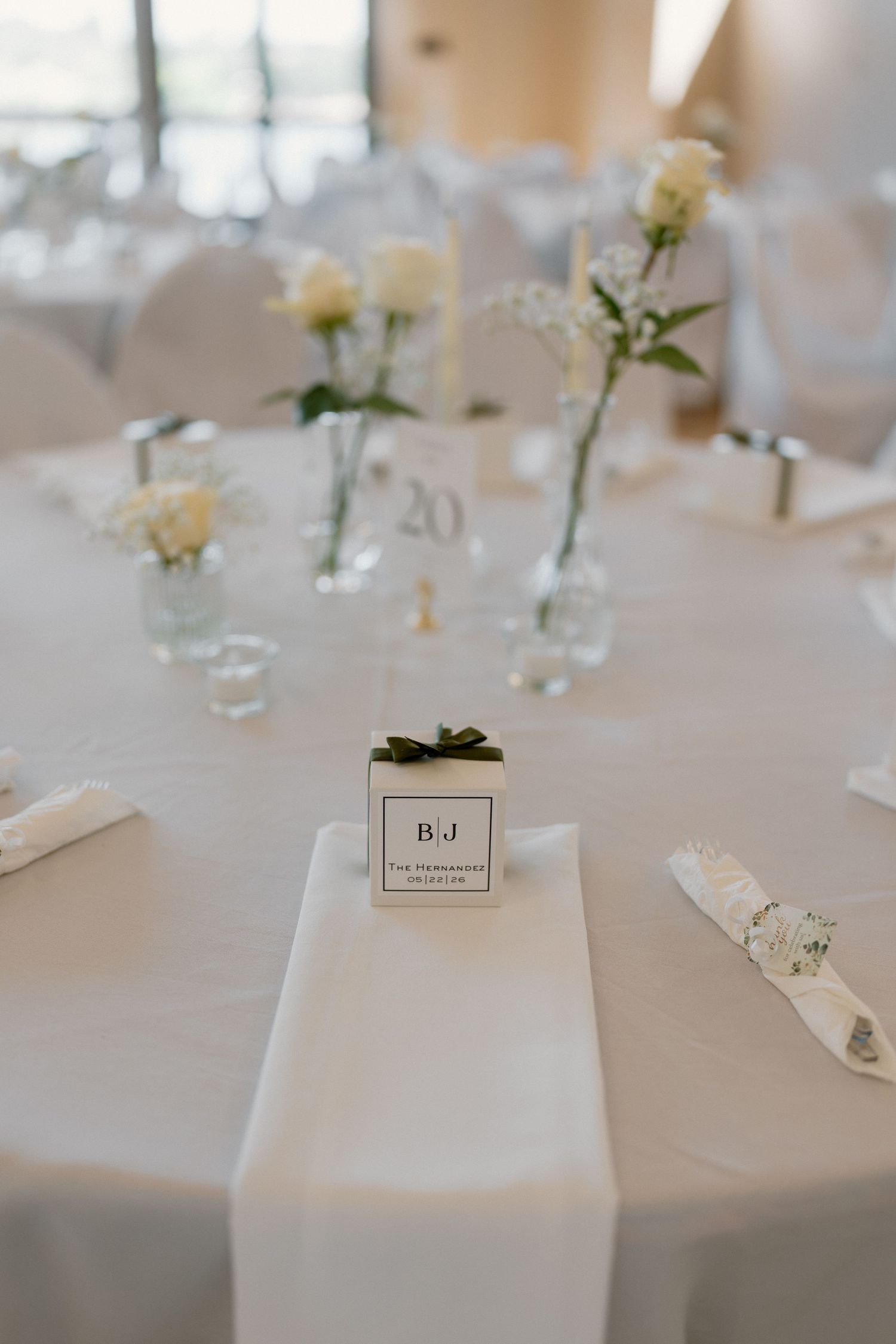 White wedding reception table setting with monogrammed favor box, white roses, and elegant decor.