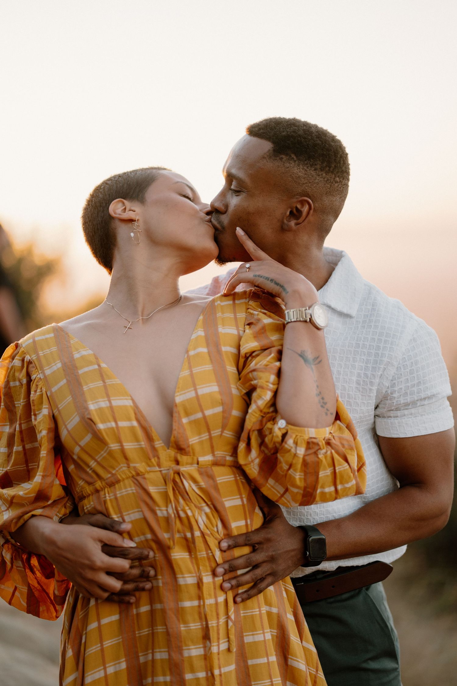 A couple shares a passionate kiss at sunset during an outdoor engagement photo session.