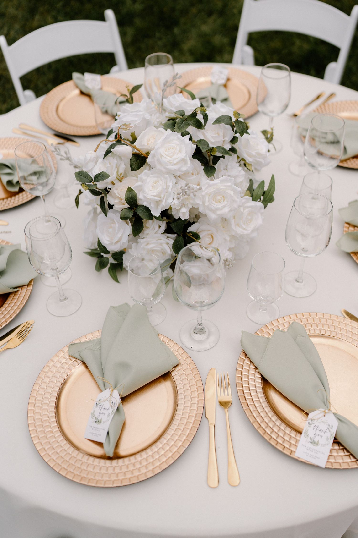 Elegant wedding reception table with gold charger plates, sage green napkins, and white rose floral centerpiece.