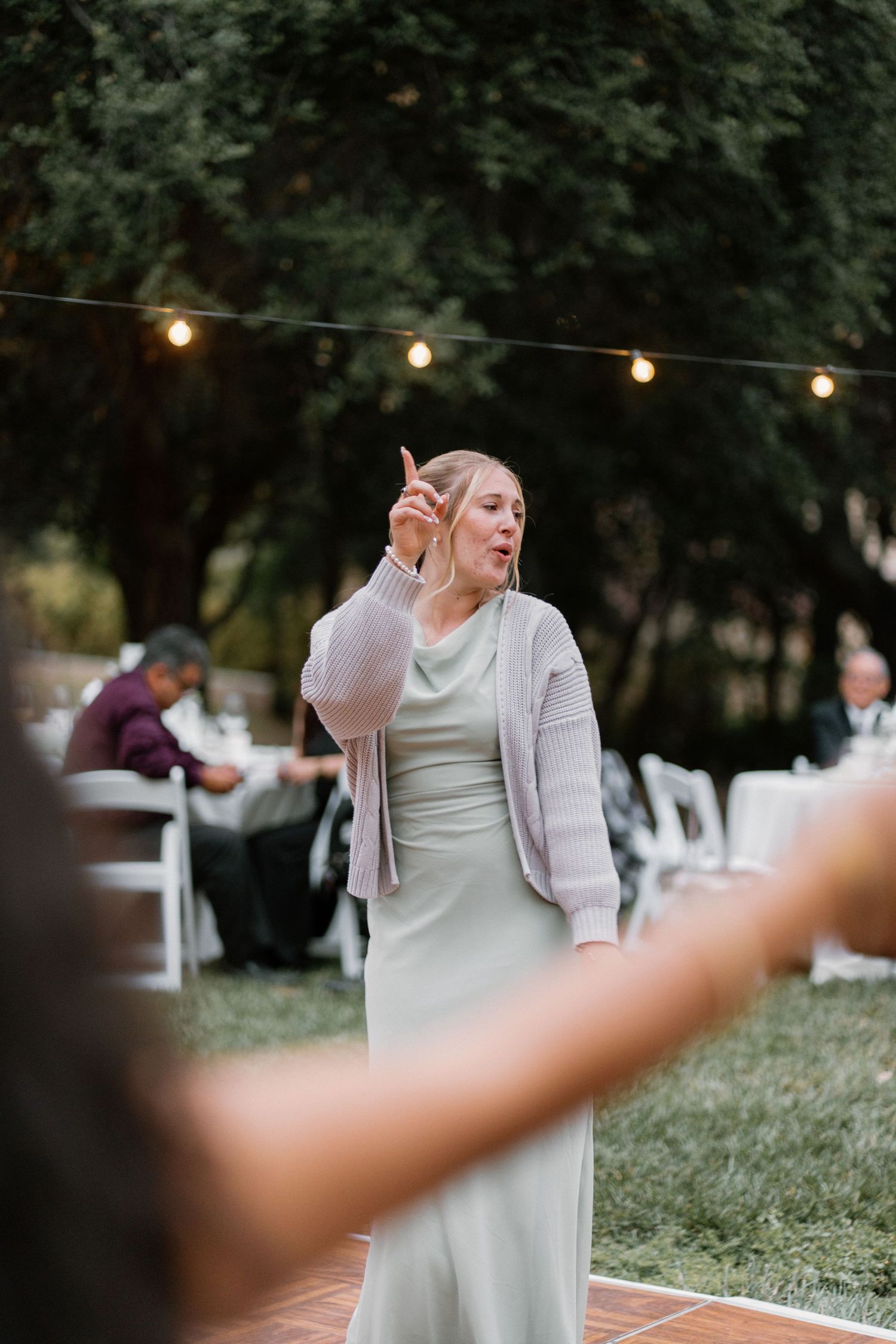A woman in a sage green dress and pink cardigan dances outdoors at a wedding reception under string lights.