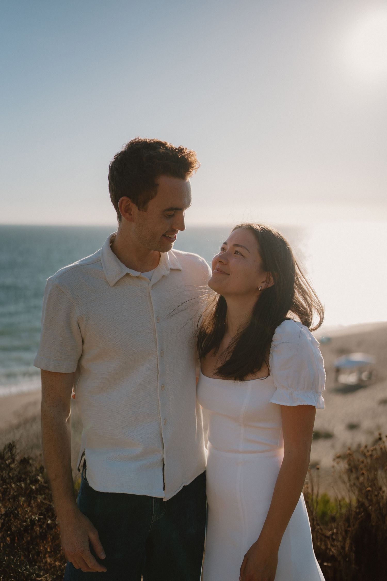 Happy couple in white outfits sharing a romantic moment at a scenic ocean overlook during sunset.
