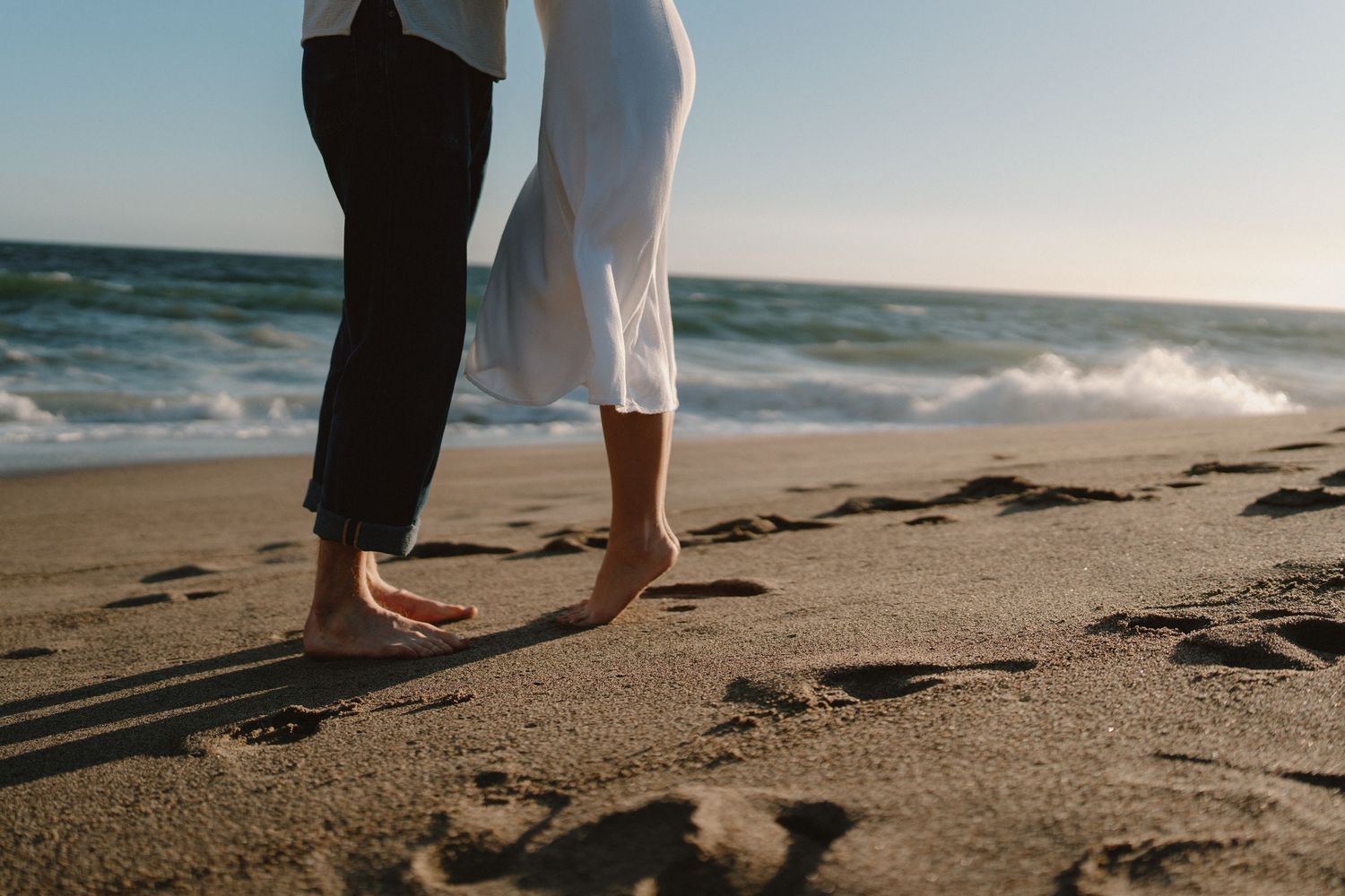 Barefoot couple standing close together on sandy beach with ocean waves in background.