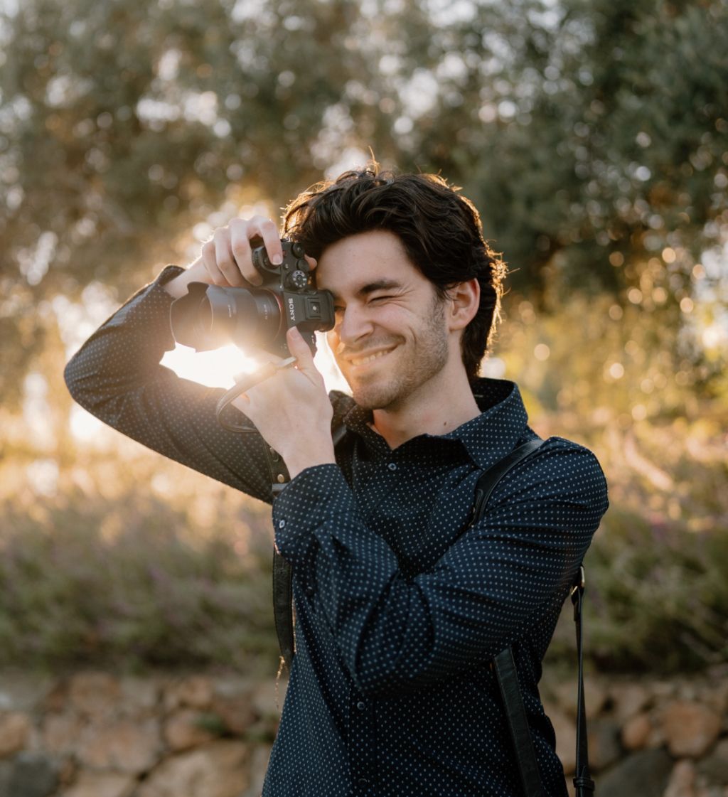 Young man smiling while photographing with a camera outdoors in golden hour sunlight among trees.