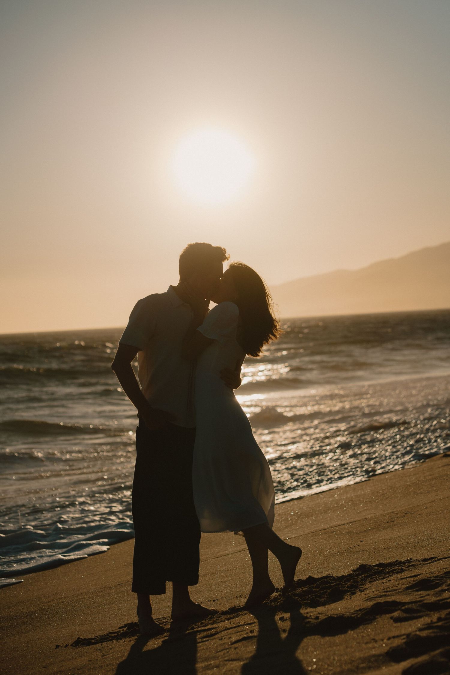 Silhouette of a romantic couple kissing on a sandy beach at golden sunset with ocean waves and mountains in background.