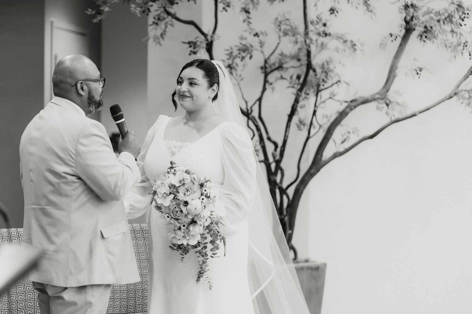 Black and white photo of a bride holding a bouquet smiling at the groom during an outdoor wedding ceremony.