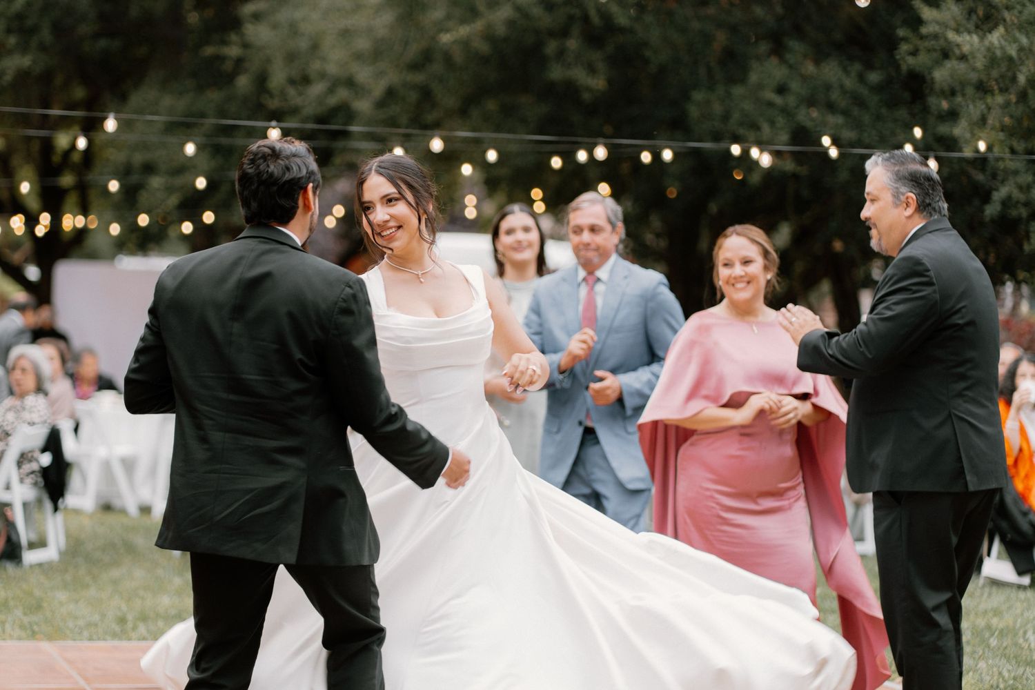 Bride in white gown dancing outdoors at wedding reception with string lights, groom in black suit, guests watching.