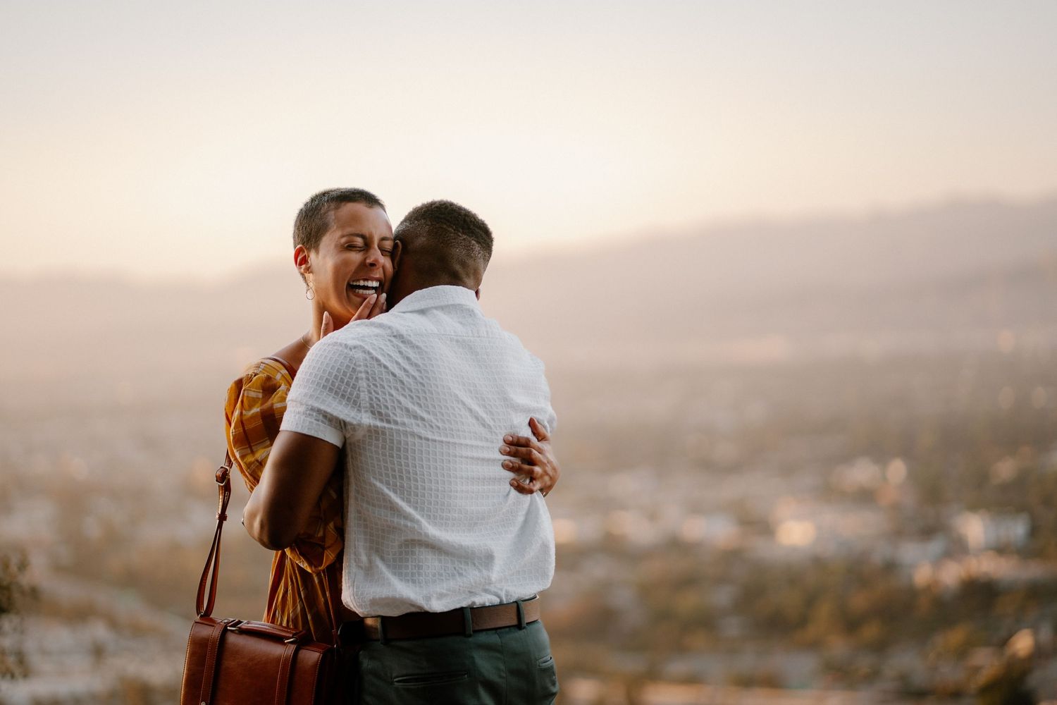 Couple embracing and laughing at sunset with city skyline in background on hilltop overlook.